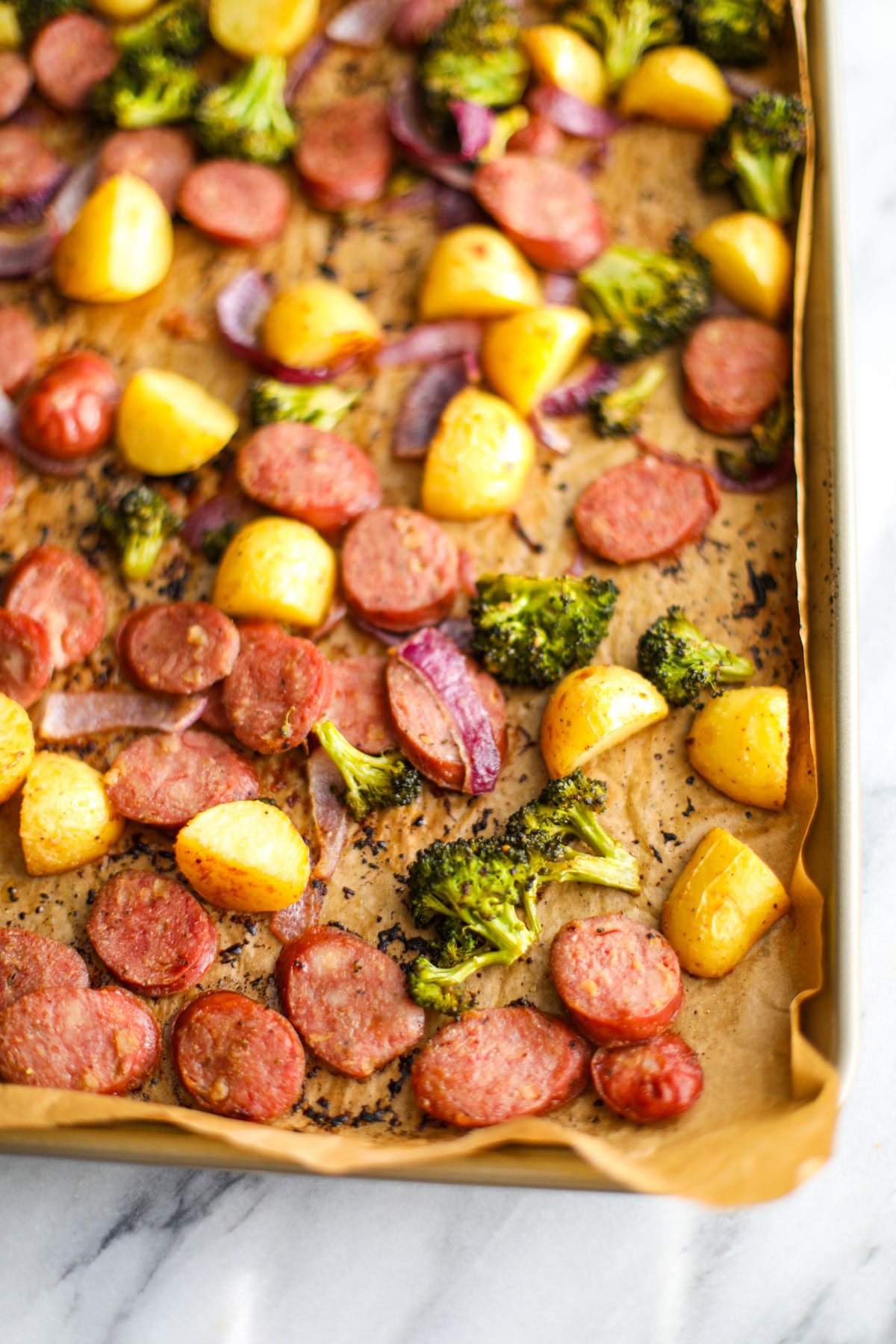 An up close of a chicken sausage sheet pan dinner with caramelized, sliced chicken sausage, baby gold potatoes, broccoli florets, and sliced red onion.