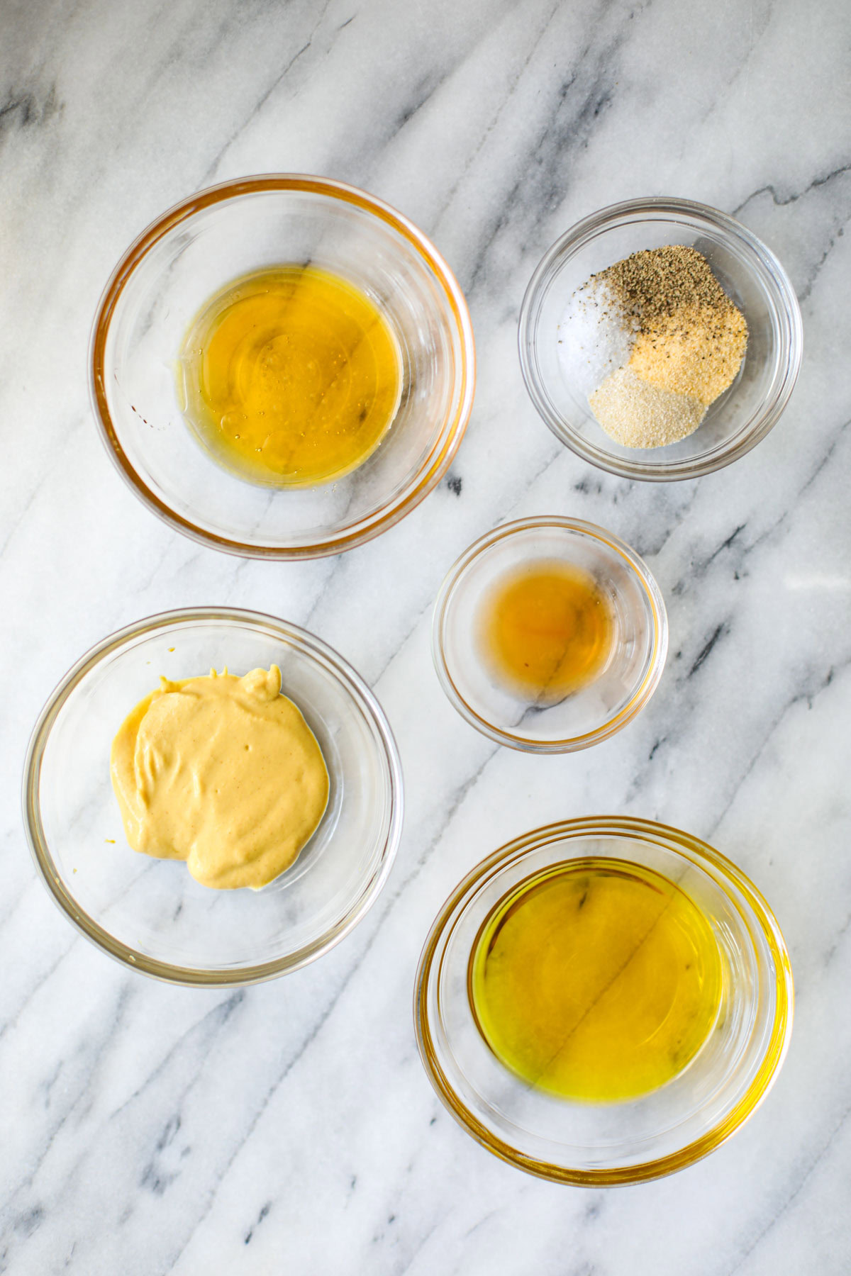 Glass bowls of ingredients to make the honey mustard dressing including honey, seasonings, vinegar, dijon mustard, and olive oil.
