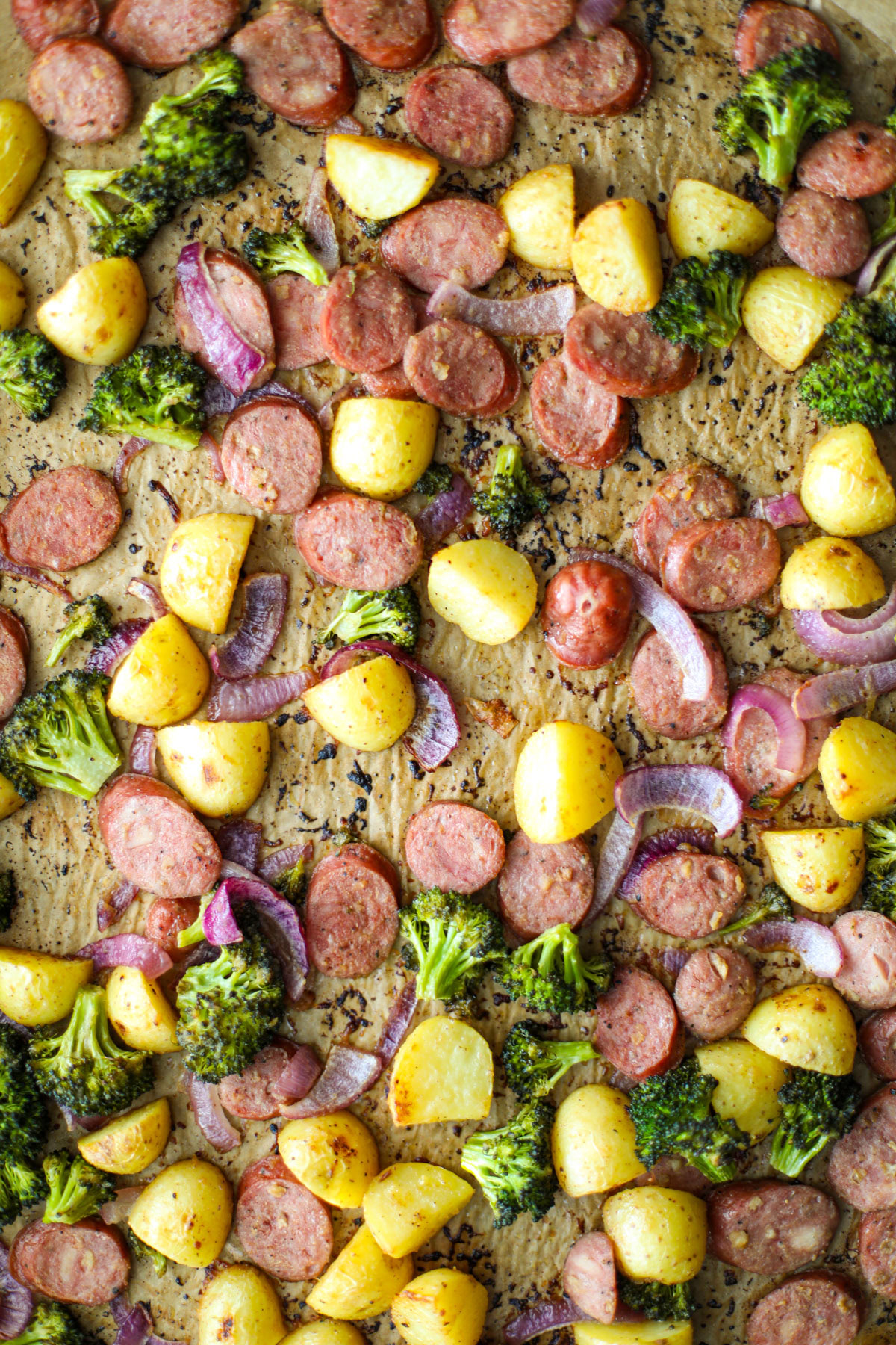 A sheet pan lined with parchment with sliced chicken sausage, baby gold potatoes, broccoli florets, and sliced red onion.