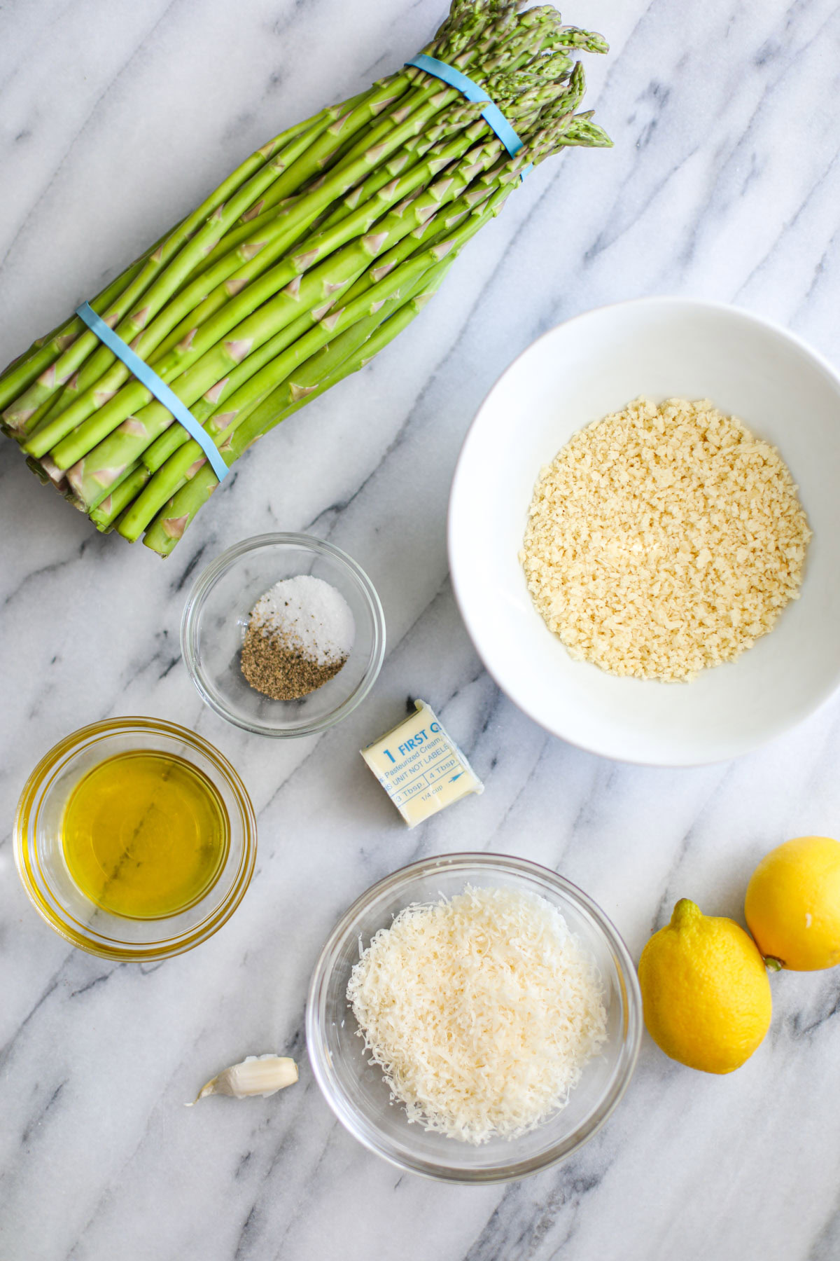 A bundle of asparagus, a white bowl of panko breadcrumbs, a glass bowl of seasonings, two tablespoons of butter, a glass bowl of olive oil and parmigiano, two lemons, and 1 garlic clove on a marble counter. 