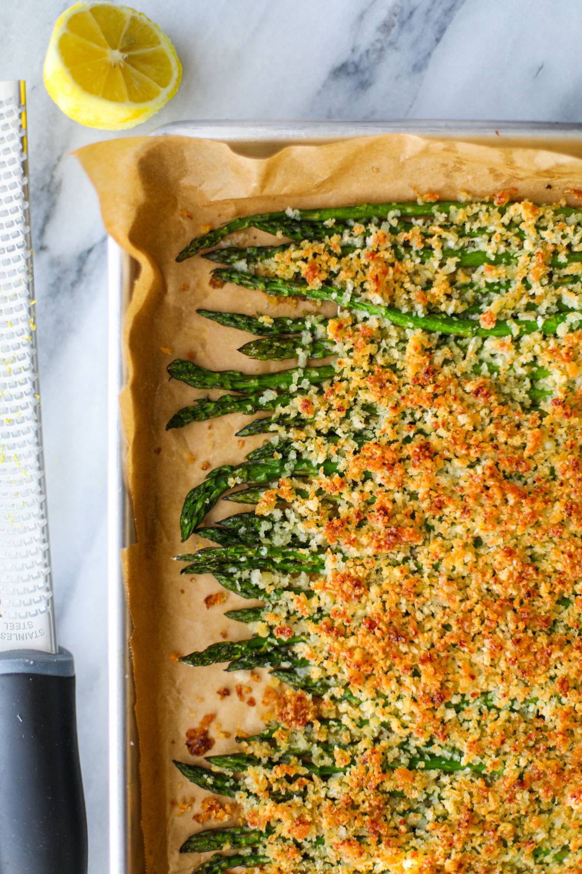 Asparagus in a single layer on a parchment lined baking sheet topped with golden breadcrumbs, with a microplane, and lemon.