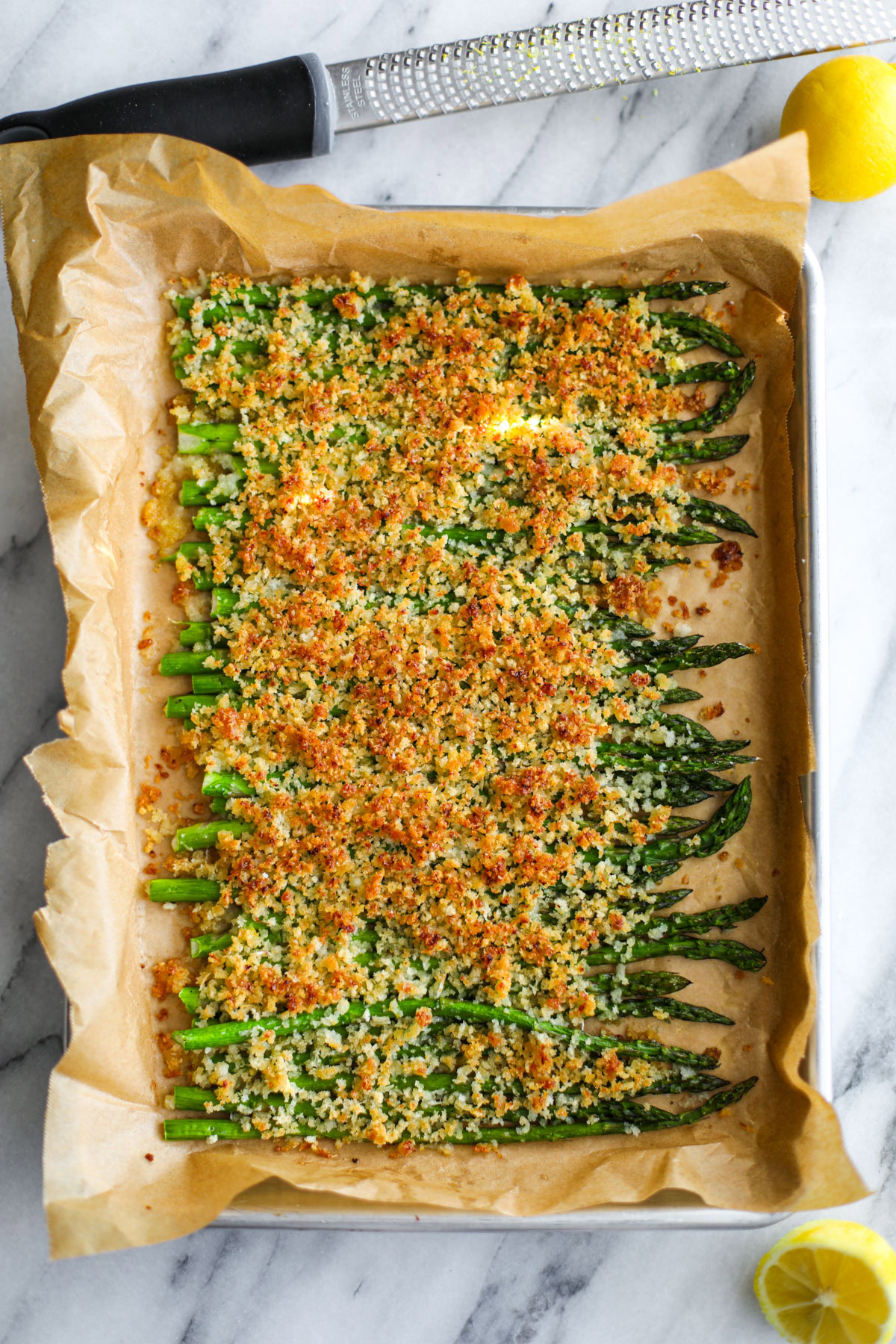 Lemon Parmesan Asparagus in a single layer on a parchment lined baking sheet topped with golden breadcrumbs, with a lemon sitting next to it.