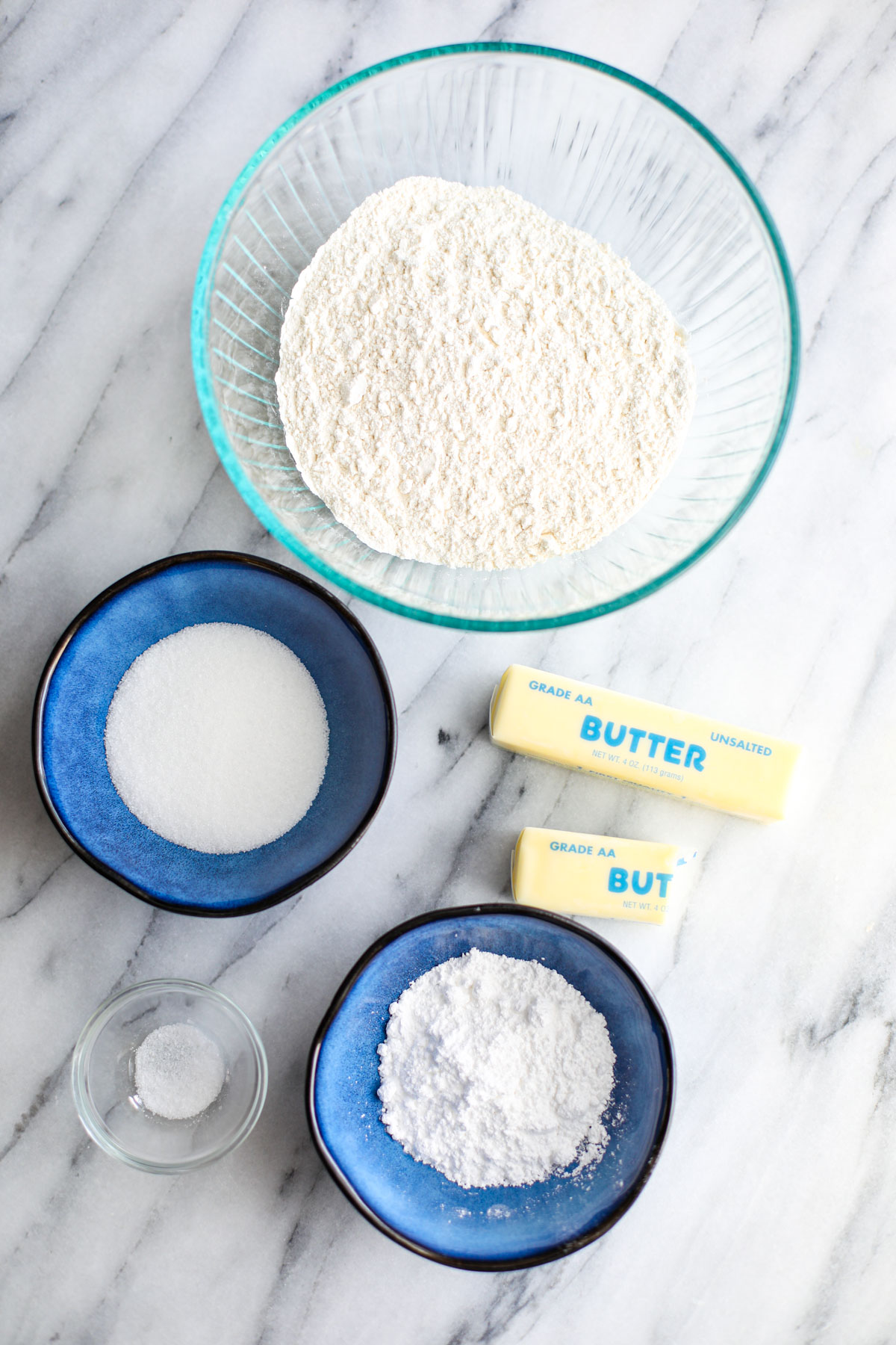 A glass bowl of flour, blue bowls of sugars, sticks of butter, and a small glass bowl of salt.