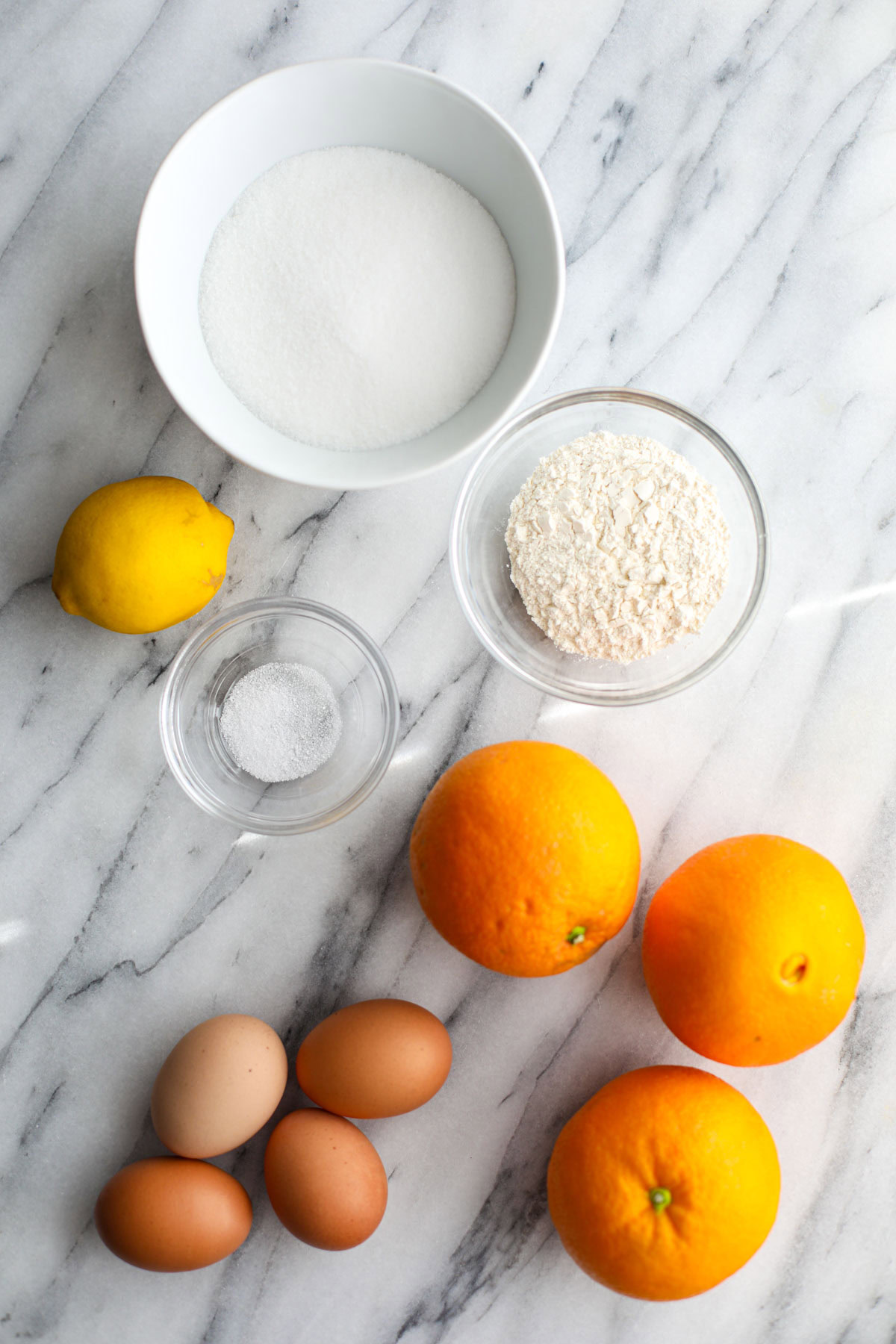 A bowl of white sugar, a lemon, three oranges, 4 eggs, salt and flour in a glass bowl.