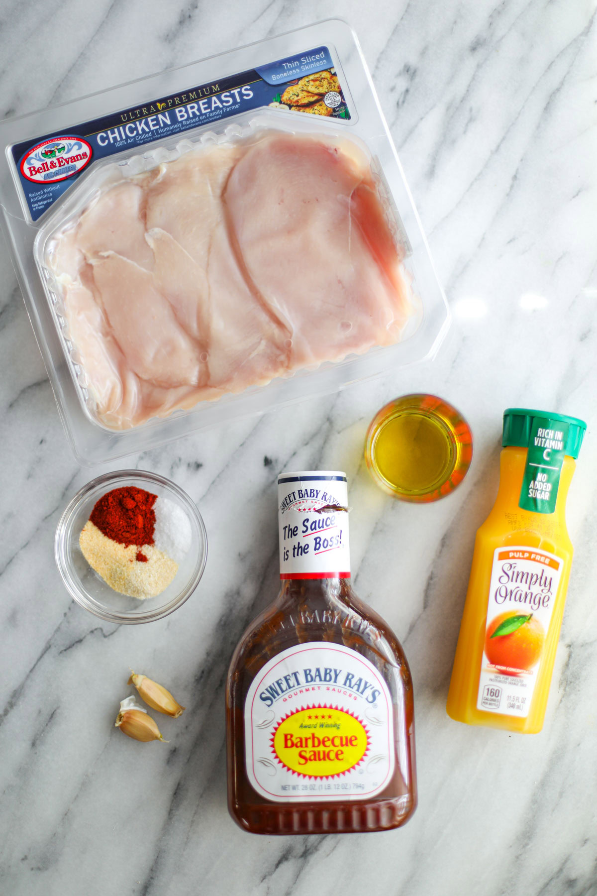 A package of chicken, a glass bowl of spices, barbecue sauce, a measuring cup of oil, and a bottle of orange juice.