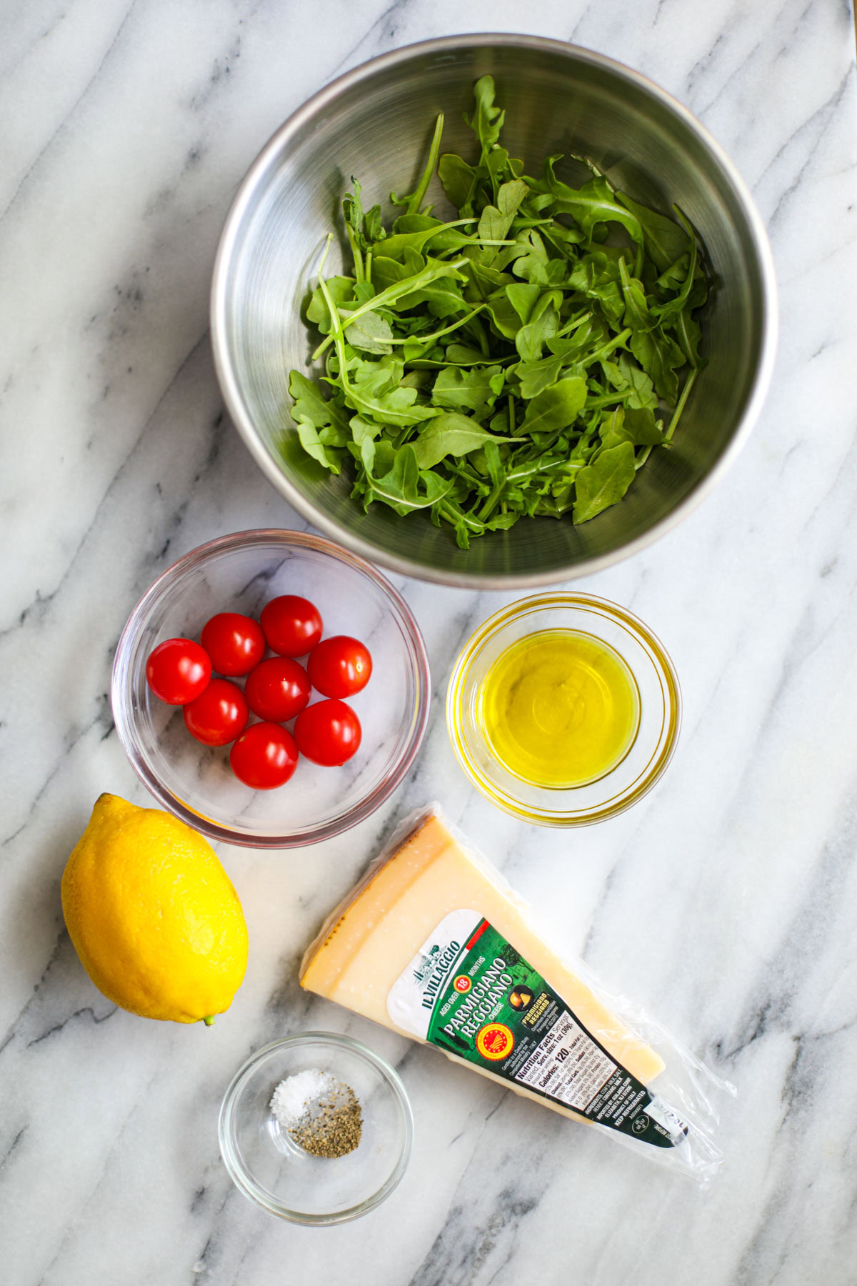 Ingredients to make an arugula side salad including arugula, cherry tomatoes, oil, lemon, parmesan, and seasonings. 