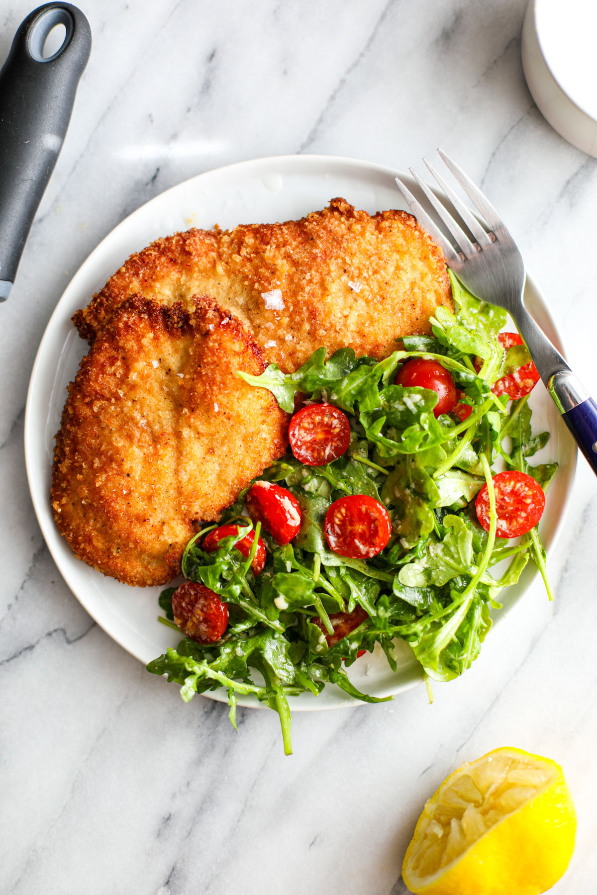 Two pieces of crispy chicken milanese with an arugula salad on a white plate with a fork on the side next to half a lemon and a microplane.