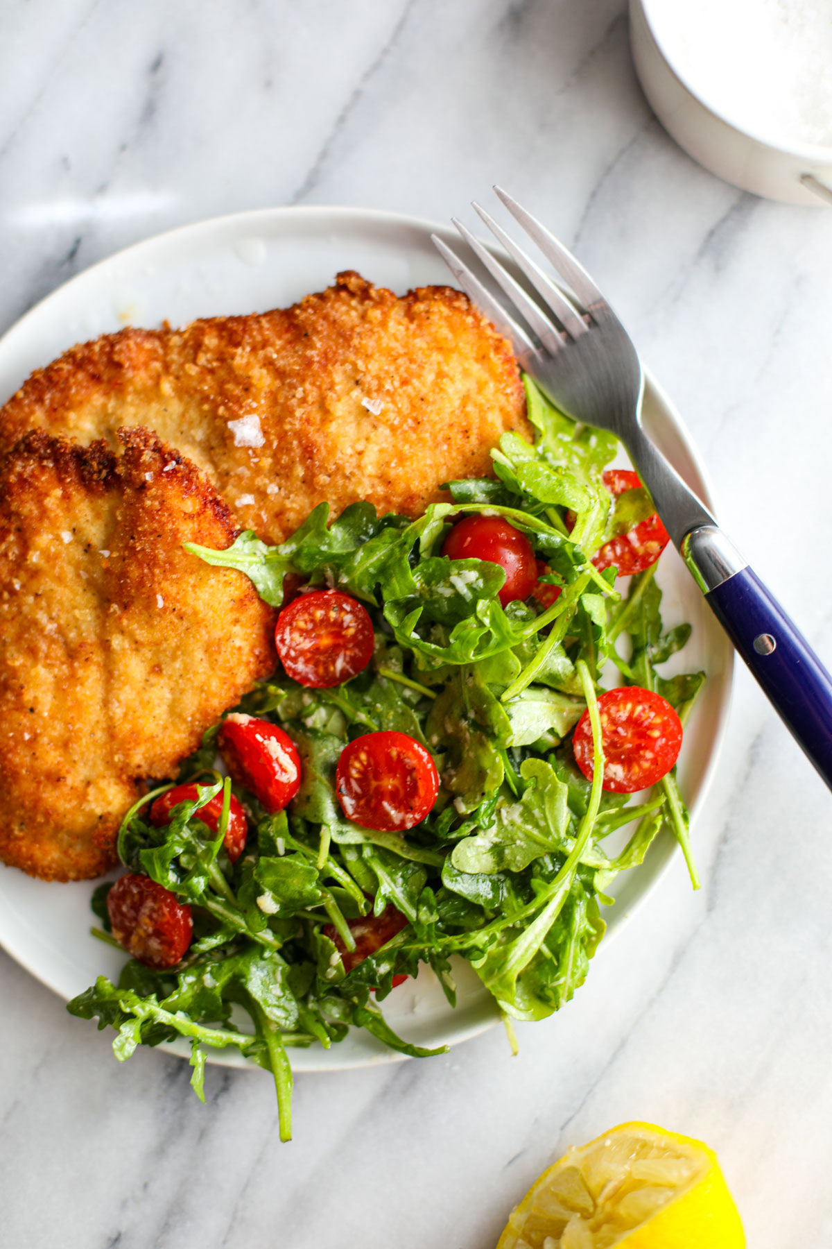 Two pieces of crispy chicken milanese with an arugula salad on a white plate with a blue fork on the side. 