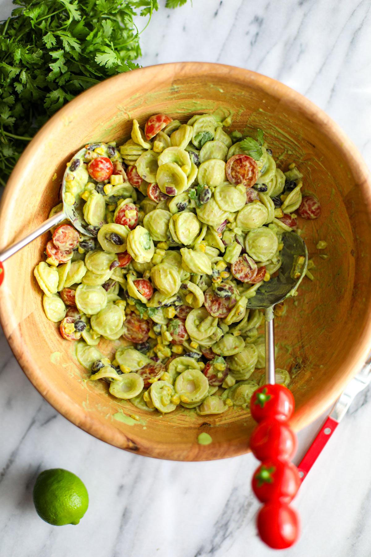 Avocado cilantro lime pasta salad in a large acacia wood bowl with red tomato salad turners.