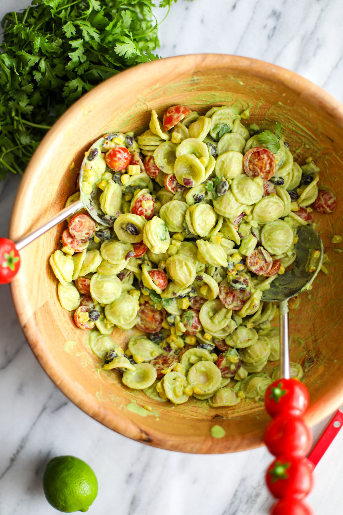 Avocado cilantro lime pasta salad in a large acacia wood bowl with red tomato salad turners.
