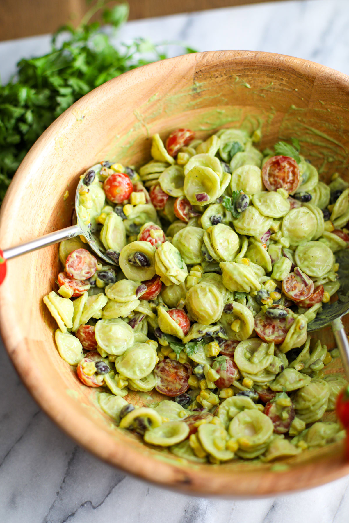 Avocado cilantro lime pasta salad in a large acacia wood bowl with red tomato salad turners.