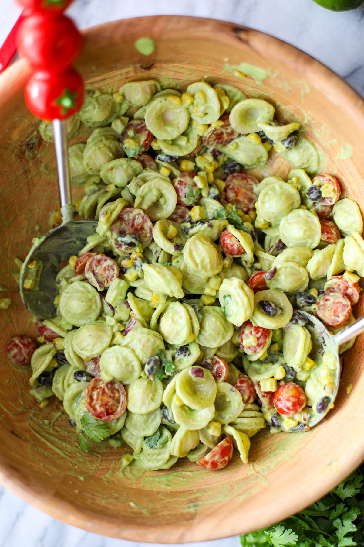 Avocado cilantro lime pasta salad in a large acacia wood bowl with red tomato salad turners.