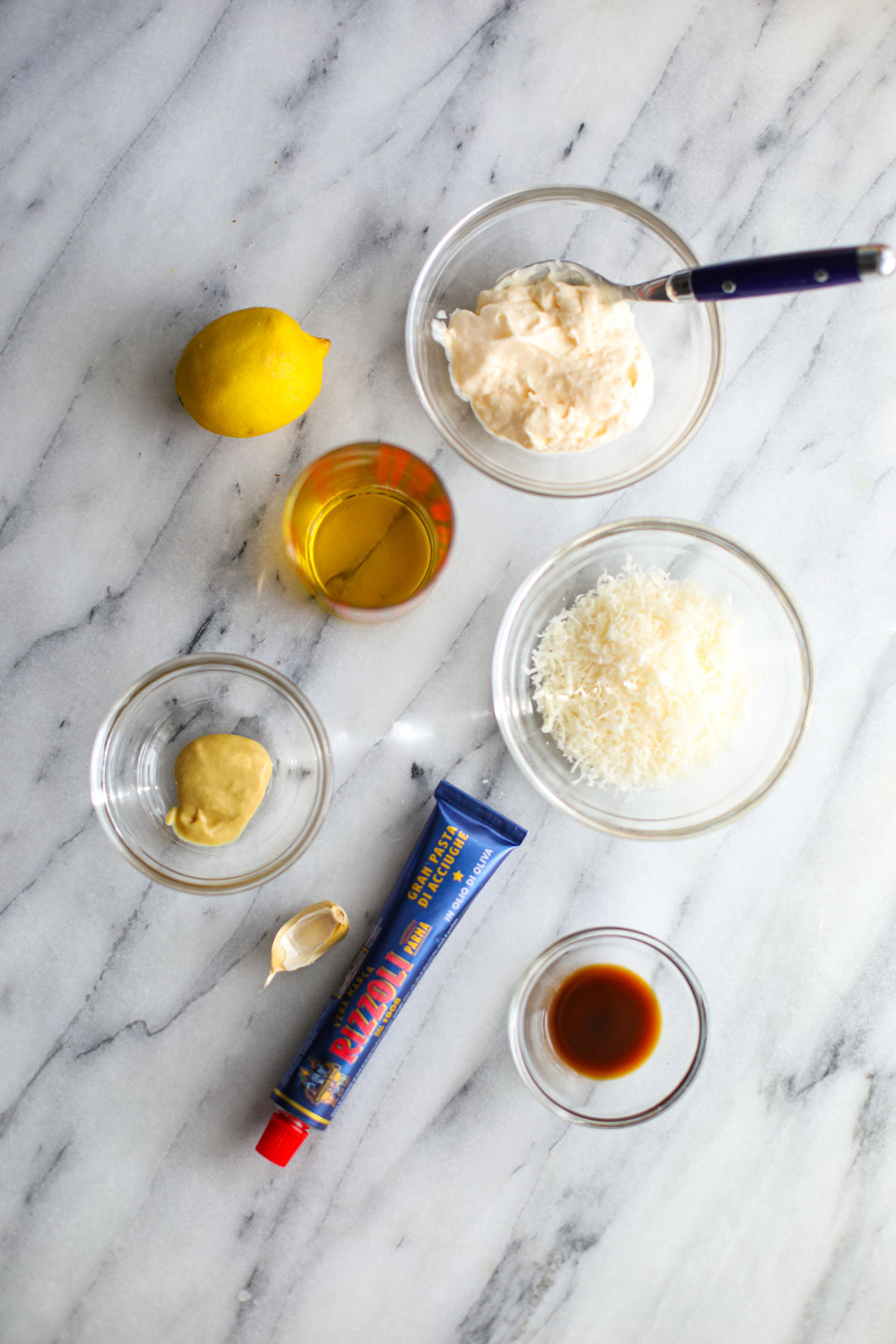 Ingredients to make homemade caesar dressing including mayonnaise, a lemon, olive oil, parmigiano reggiano, dijon mustard, Worcestershire, anchovy paste tube, and a garlic clove.
