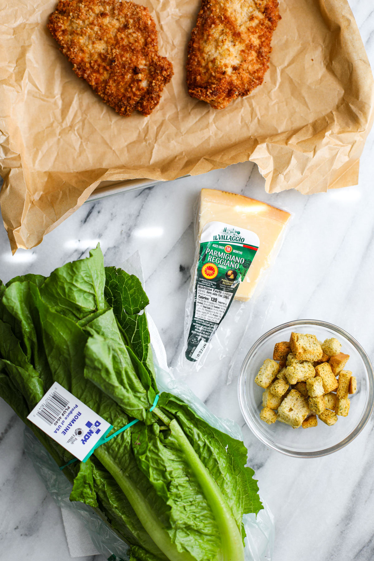 Salad ingredients including a head of romaine lettuce, two crispy chicken pieces, parmigiano reggiano wedge, and a container of croutons.