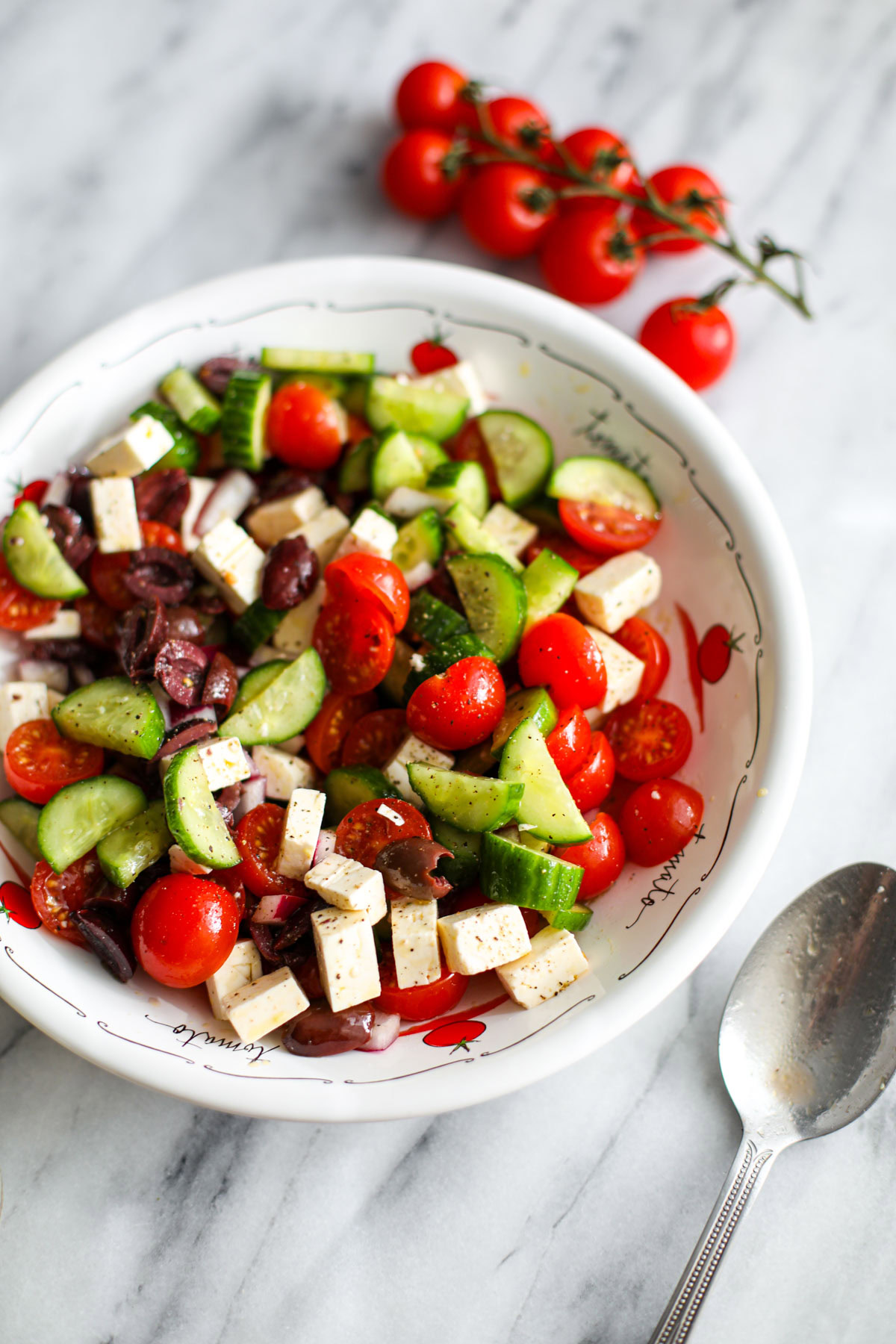 A white low profile bowl with cucumber tomato feta salad and a silver spoon on the counter.