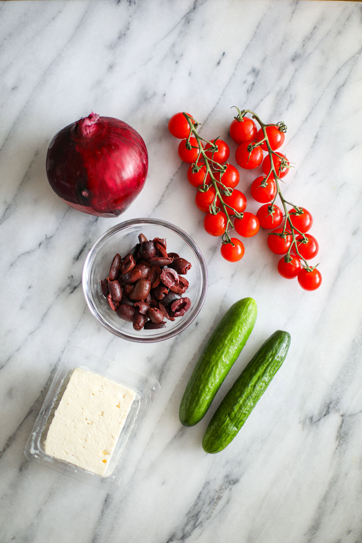 Ingredients for the salad including a large red onion, cherry tomatoes on the vine, a bowl of sliced Kalamata olives, a chunk of feta, and two small cucumbers. 