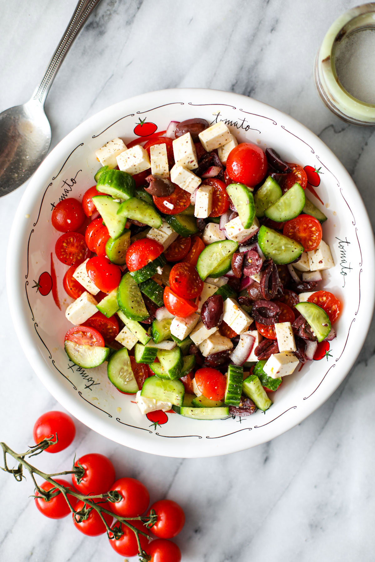 A white low profile bowl with cucumber tomato feta salad and a silver spoon on the edge and vine cherry tomatoes off to the side. 
