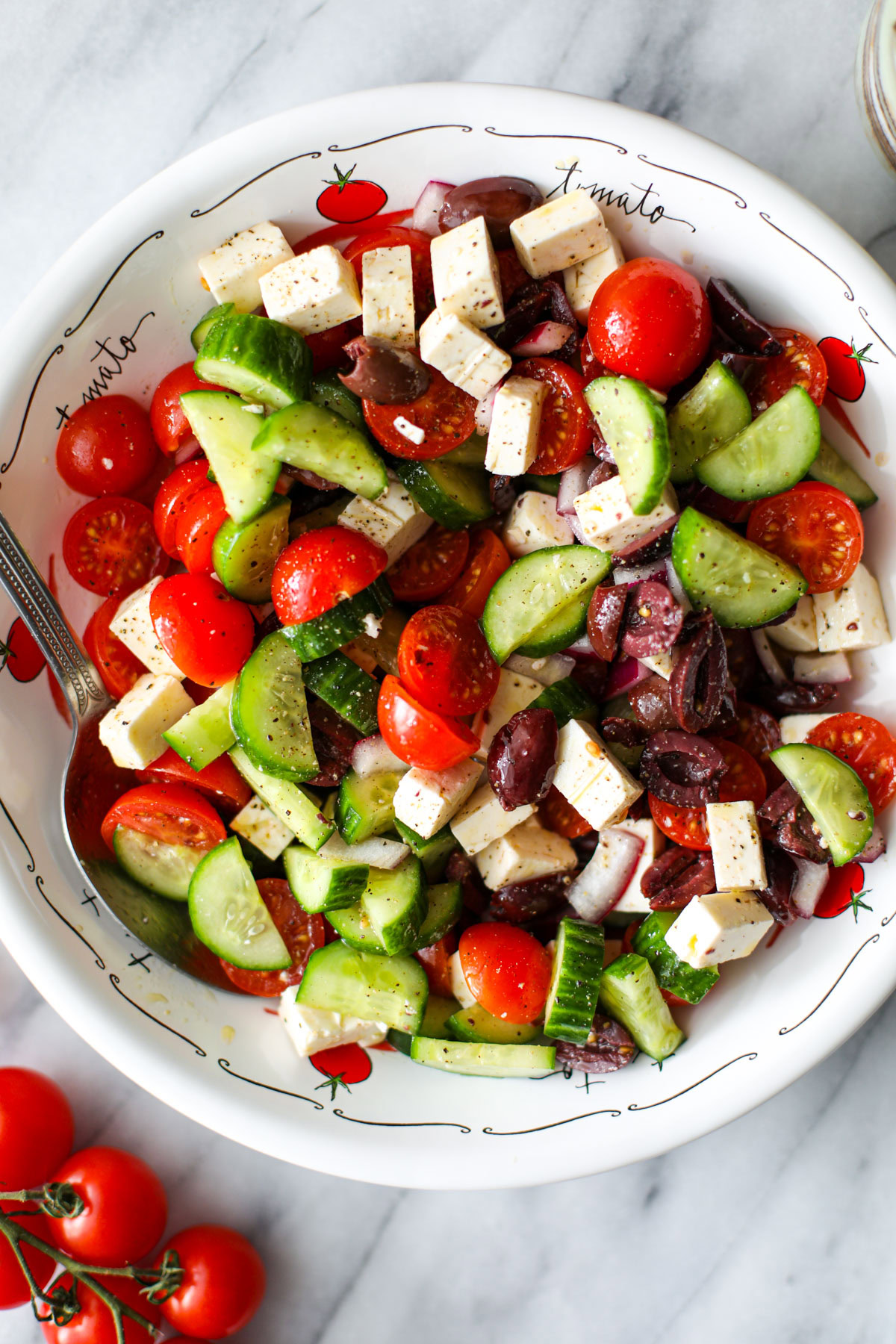 A white low profile bowl with cucumber tomato feta salad and a silver spoon on the edge. 