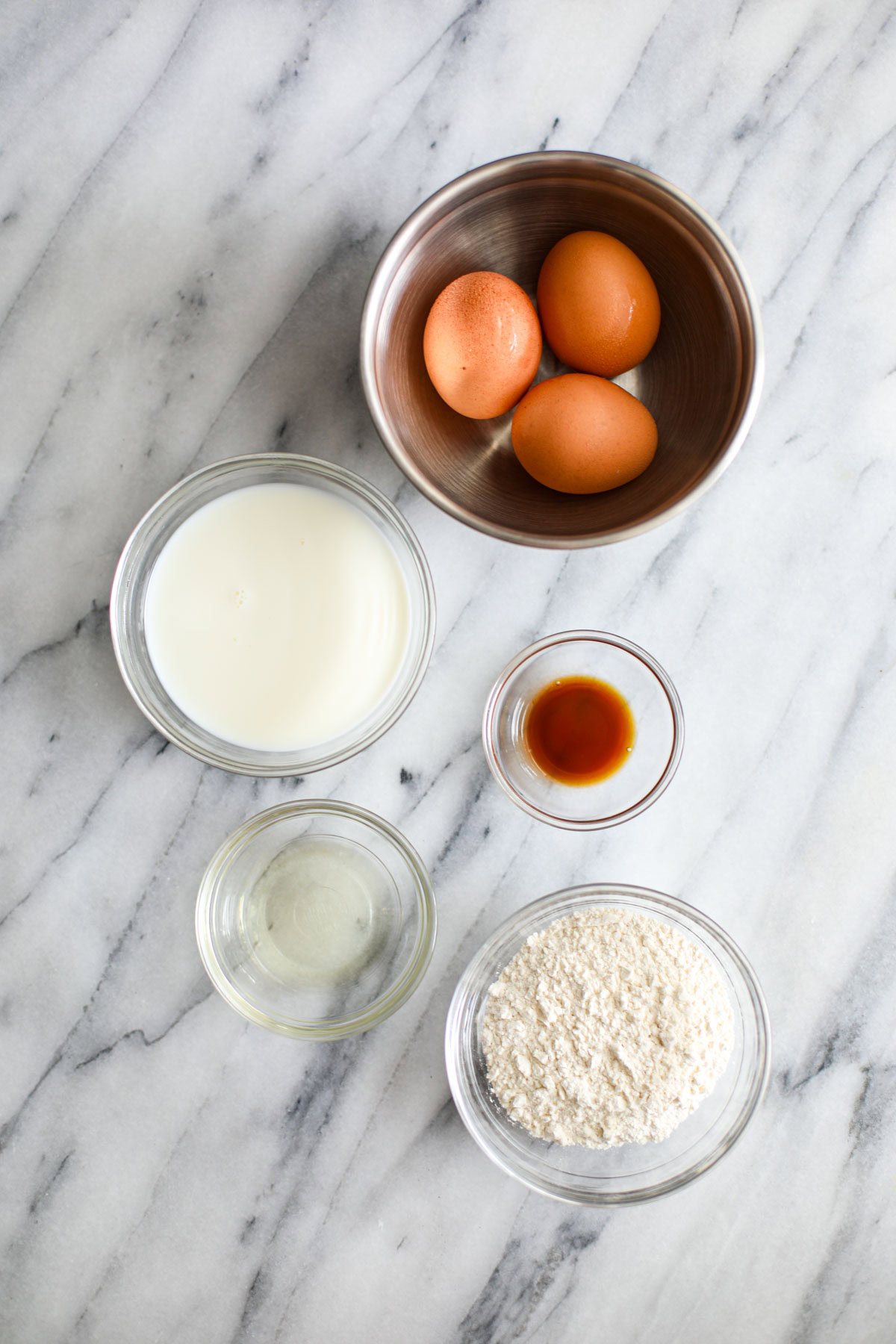 Ingredients to make a dutch baby recipe including eggs in a stainless steel bowl and glass bowls of milk, flour, vanilla, and oil. 
