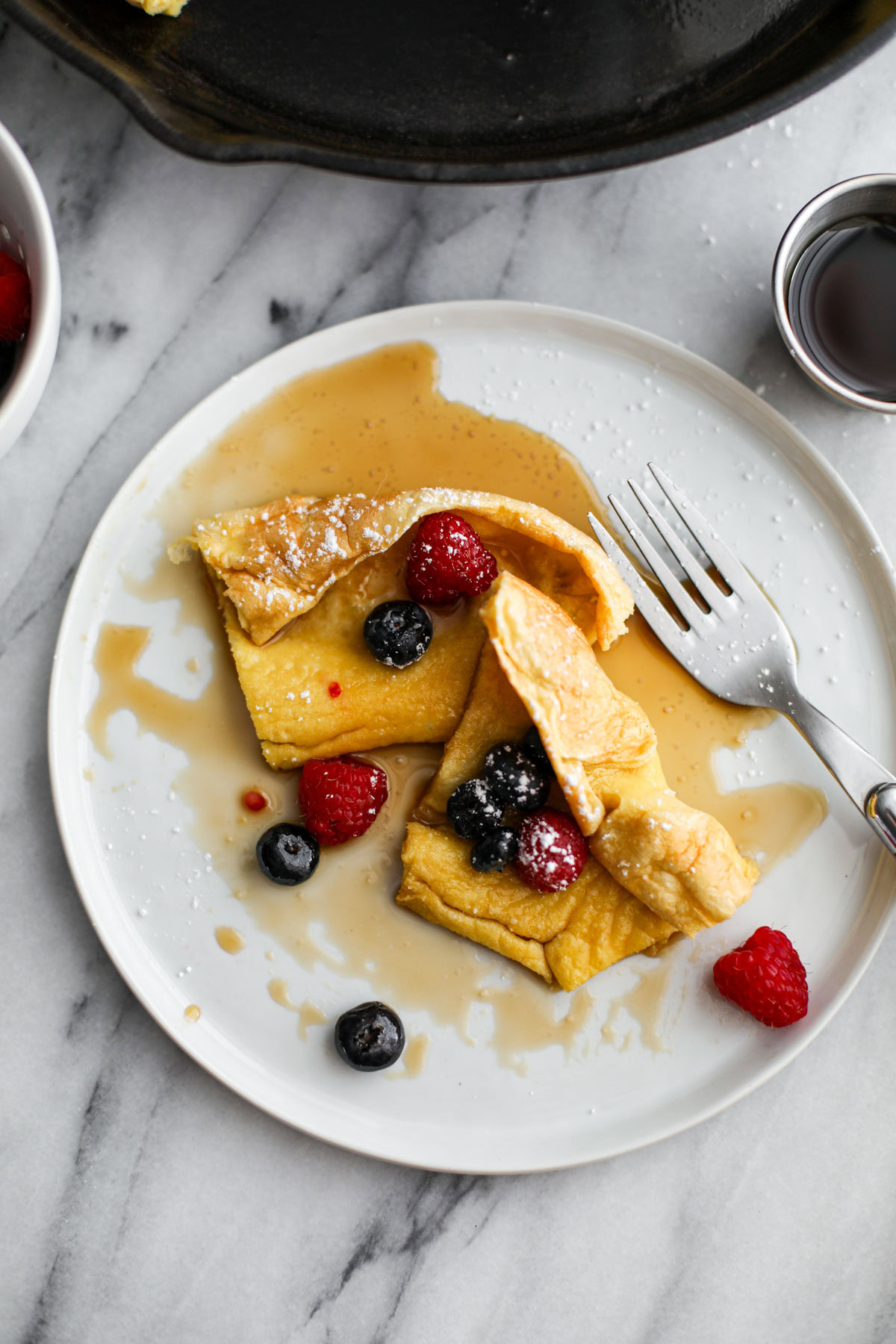 A white plate with a slice of dutch baby with maple syrup and a fork on the side with a silver ramekin to the side of syrup.