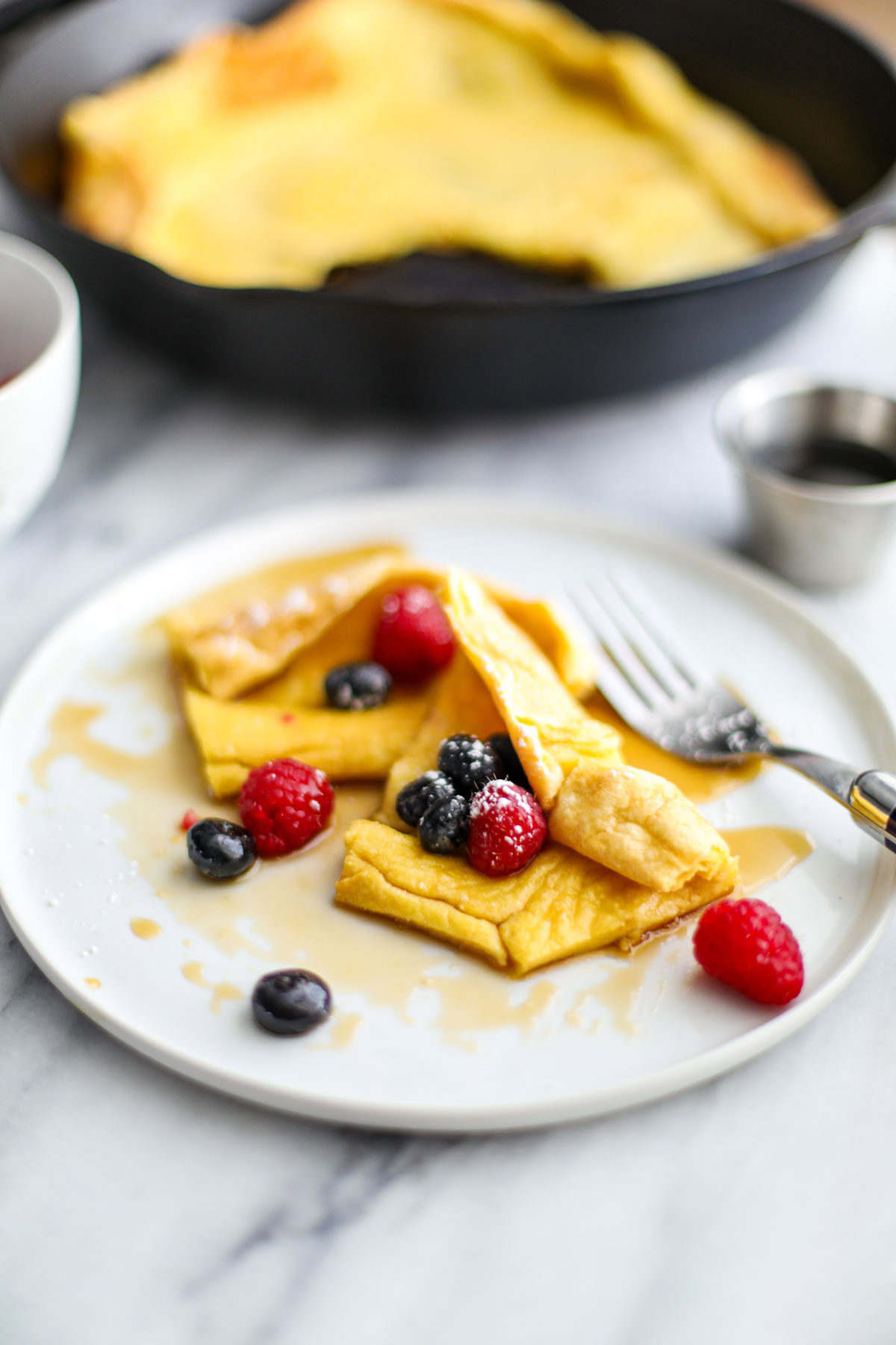 A white plate with a slice of dutch baby with maple syrup and berries and a fork on the side. 