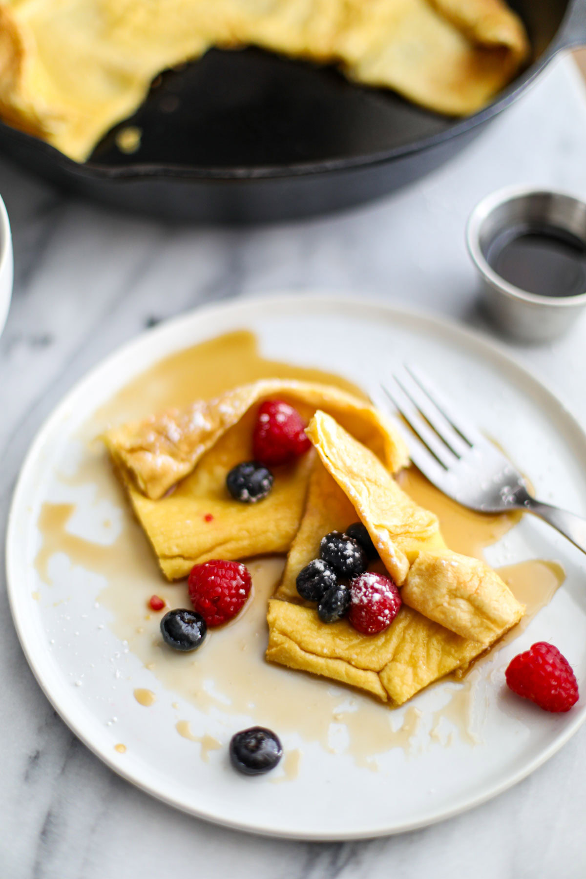 A white plate with a slice of dutch baby with maple syrup and a fork on the side. 