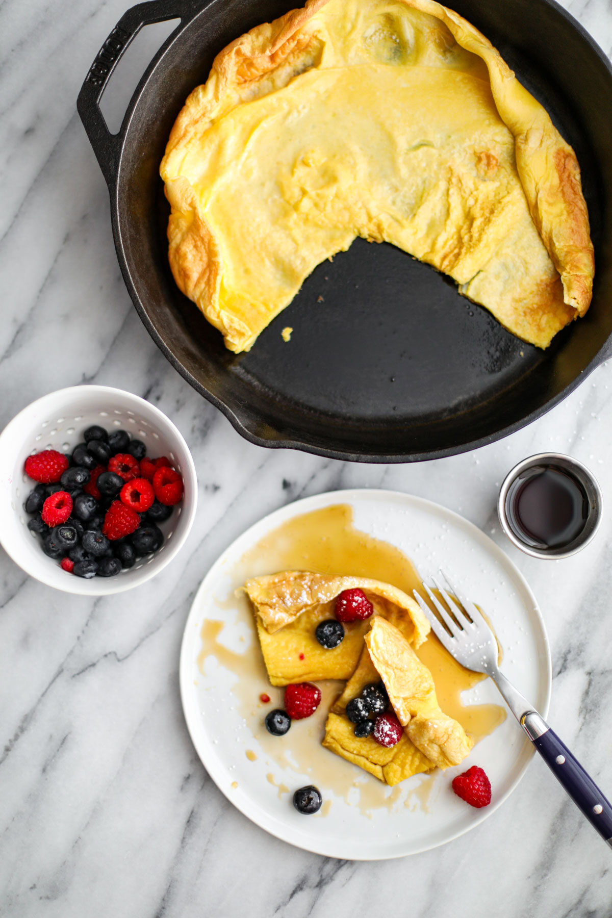 A dutch baby in a cast iron pan with a white plate with a slice on it topped with berries. 