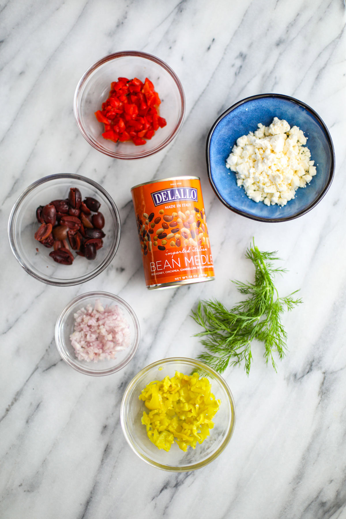 A can of beans, glass bowls of olives, shallot, pepperoncini, red peppers and a blue bowl of feta and dill.