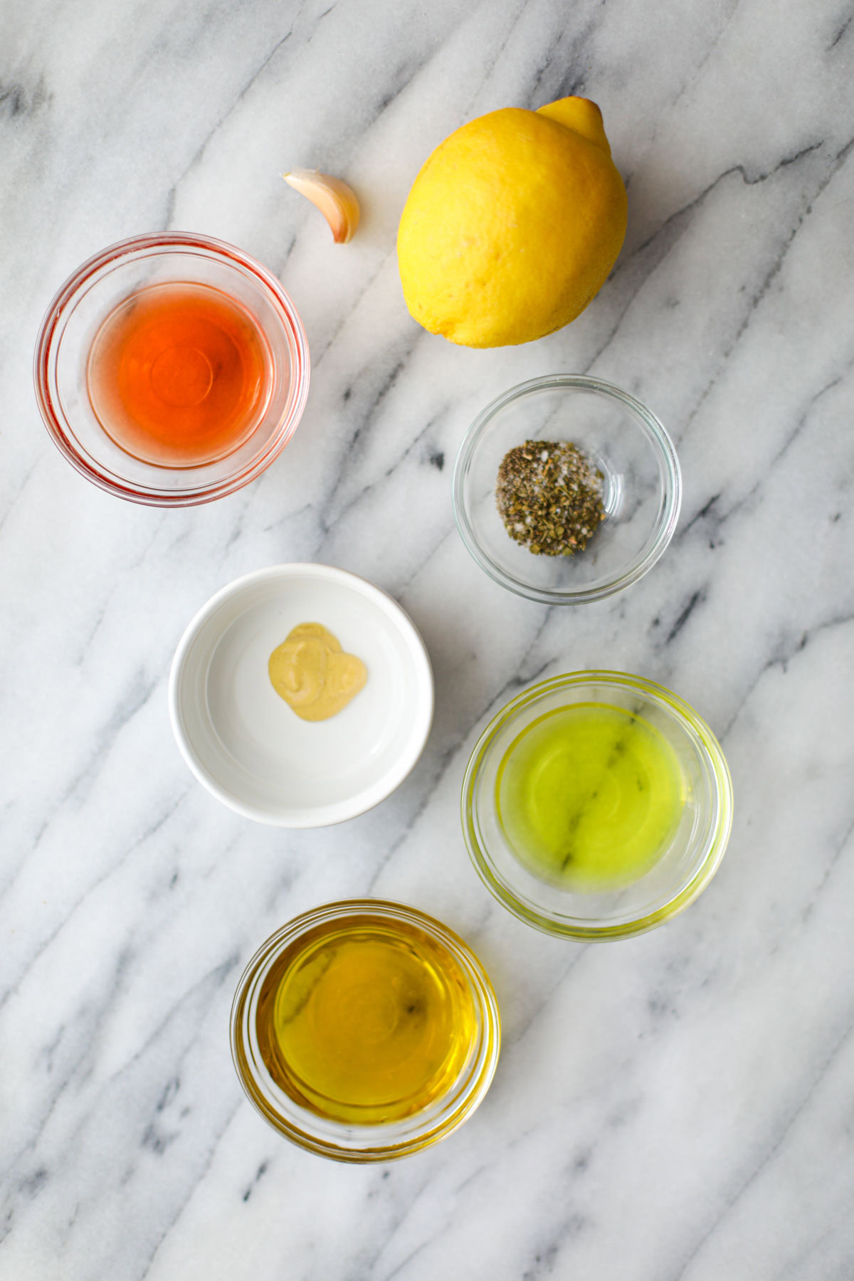 Glass bowls of red vinegar, pepperoncini brine, oregano, a whole lemon, olive oil, and a white bowl of dijon mustard.