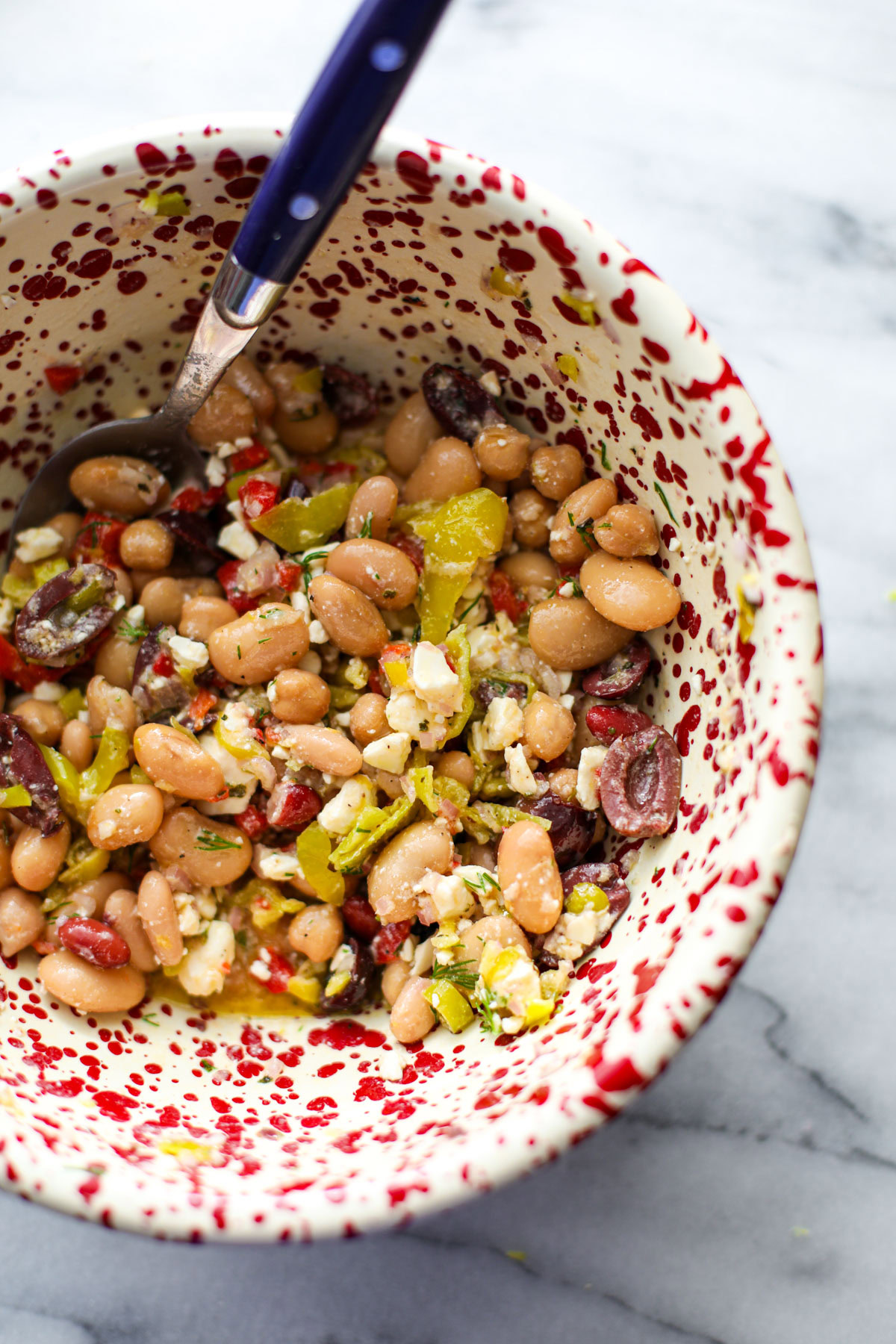 A Mediterranean dense bean salad in a red and white bowl with a blue spoon.