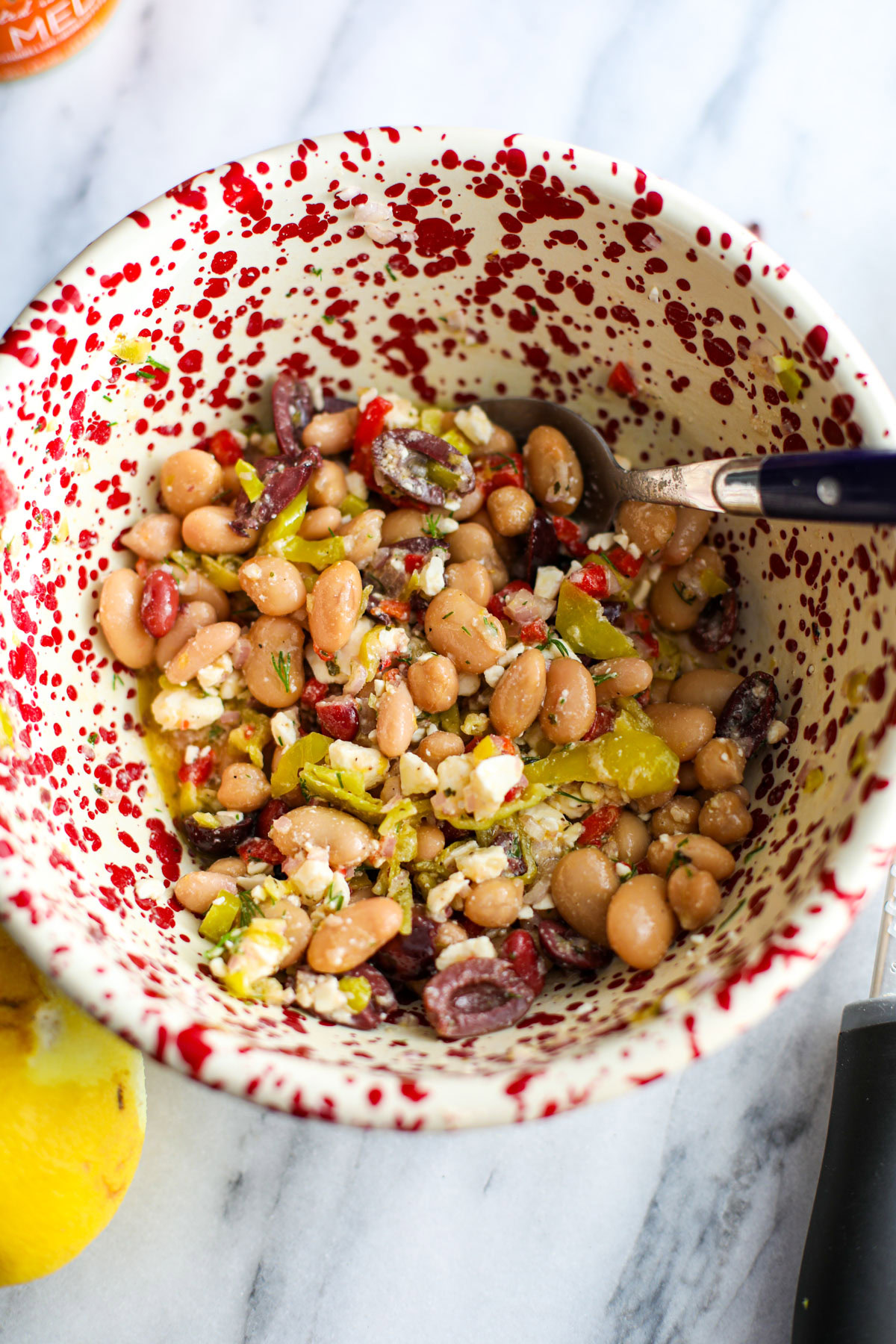 A Mediterranean dense bean salad in a red and white bowl with a blue spoon and a whole lemon.