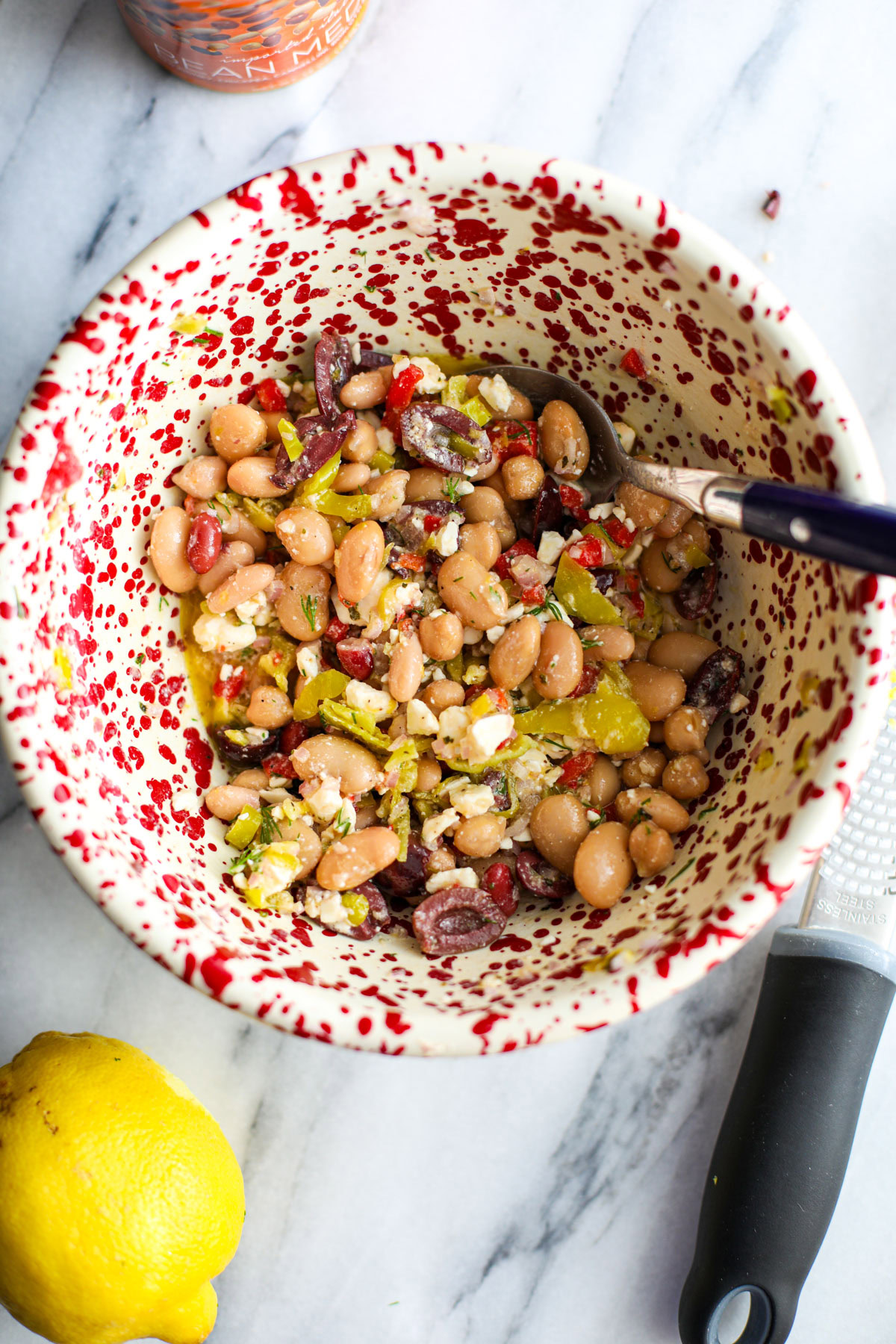 A mediterranean dense bean salad in a red and white bowl with a microplane grater and half a lemon.