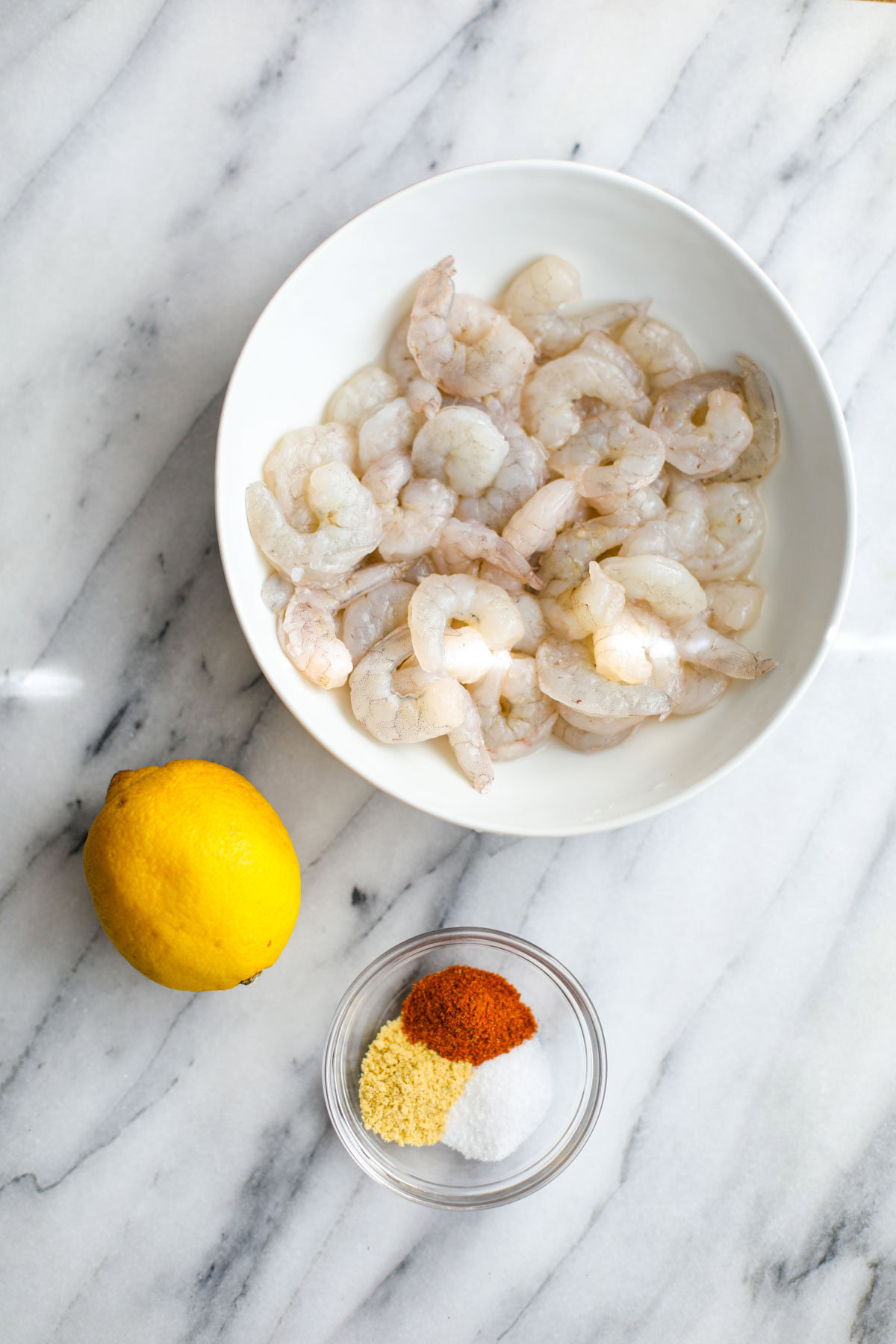 A white bowl of shrimp, a whole lemon and a glass bowl of seasonings. 