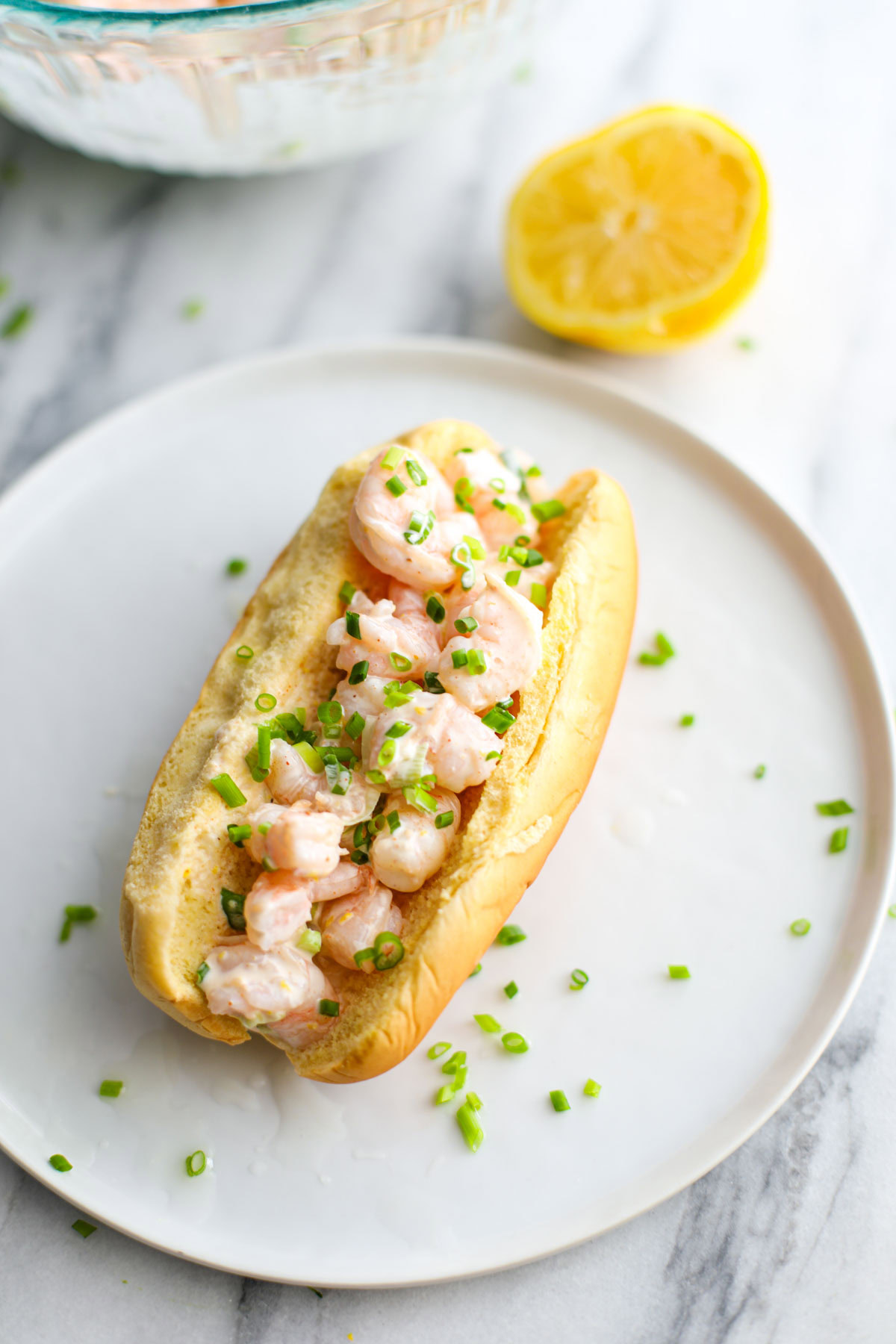 A shrimp roll topped with chives on a white plate with a lemon in the background.