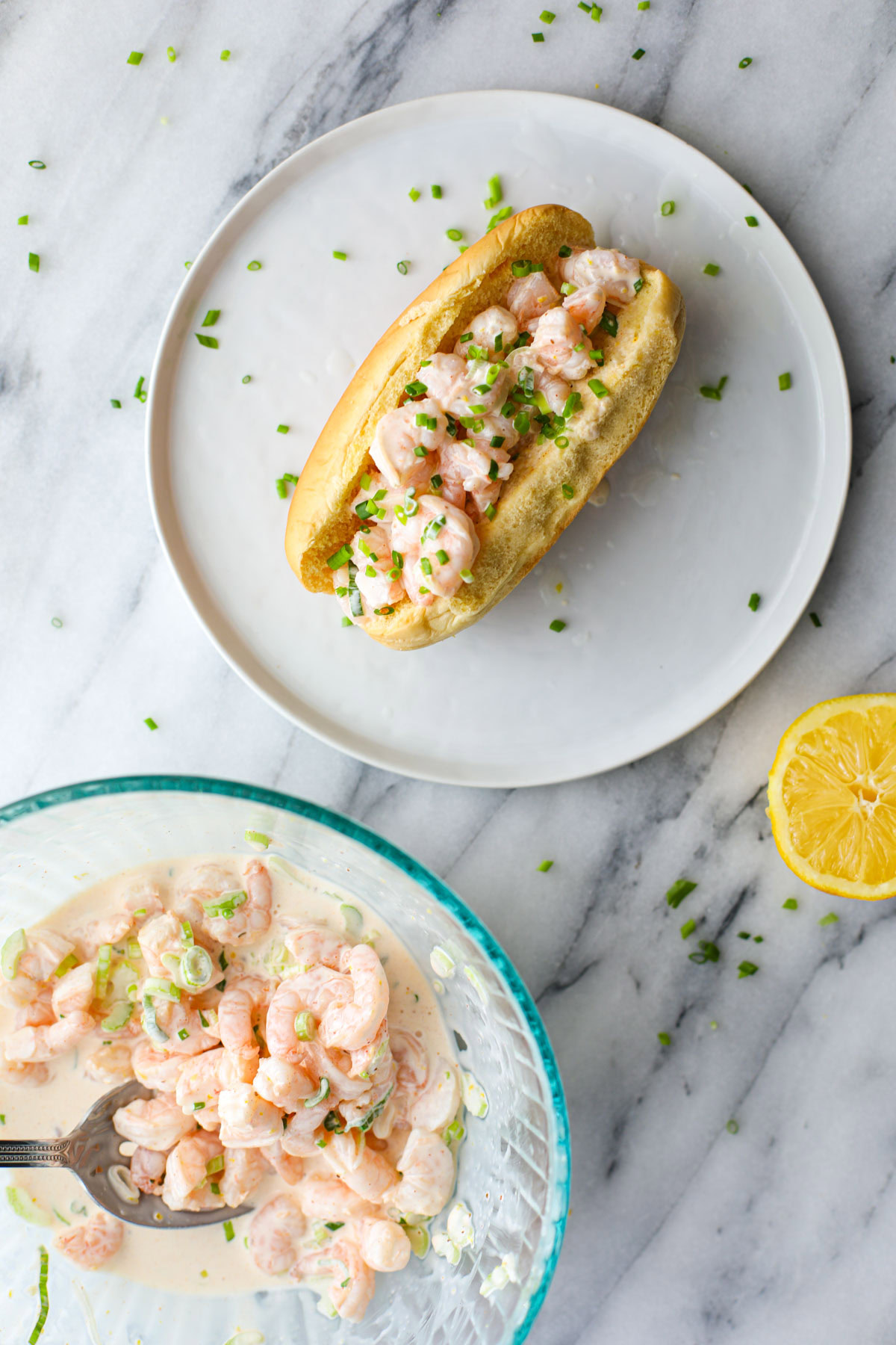 A shrimp roll topped with chives on a white plate with a glass bowl of dressed shrimp and half a lemon off to the side. 