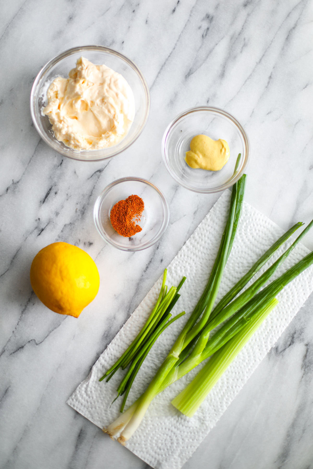 Glass bowls of mayonnaise, mustard, old bay, and a whole lemon and green onion. 