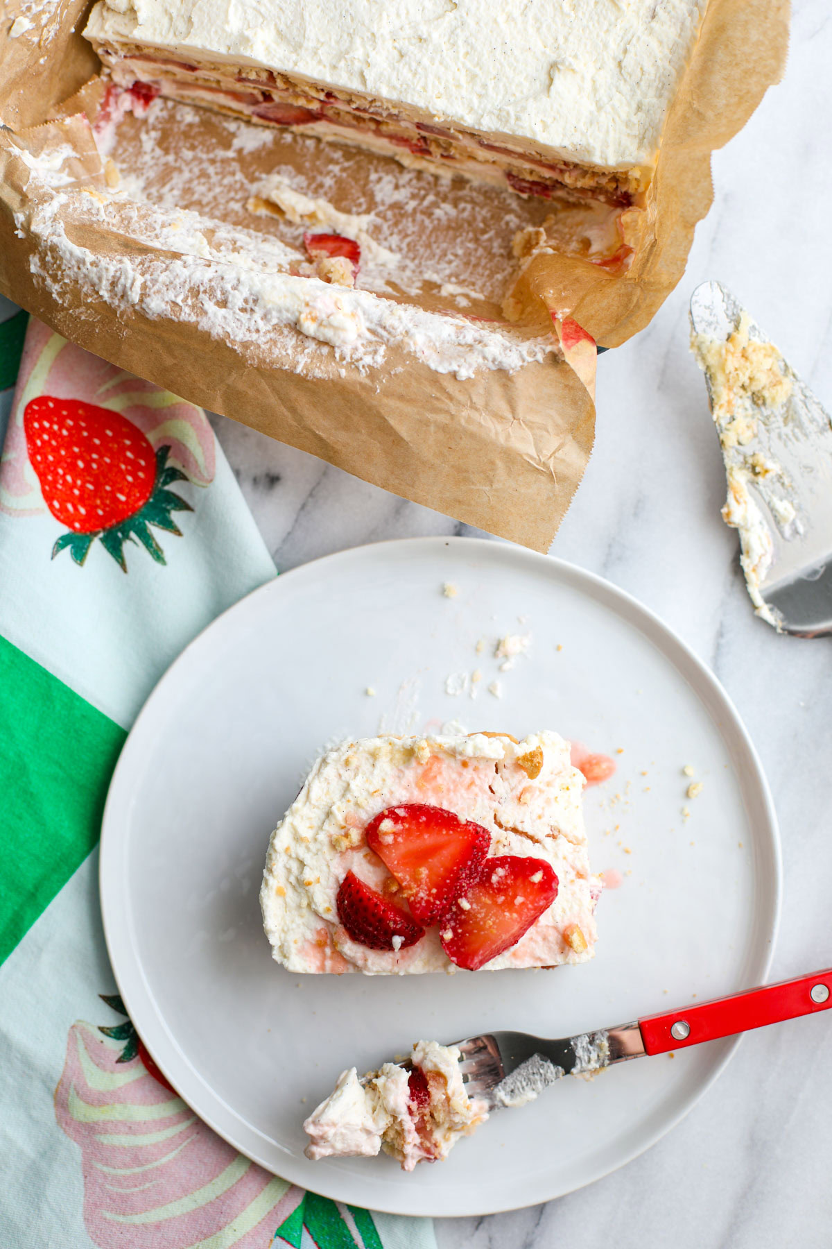 A slice of strawberry shortcake icebox cake topped with strawberries and wafer crumbs on a white plate with a red fork and pie server in the background.