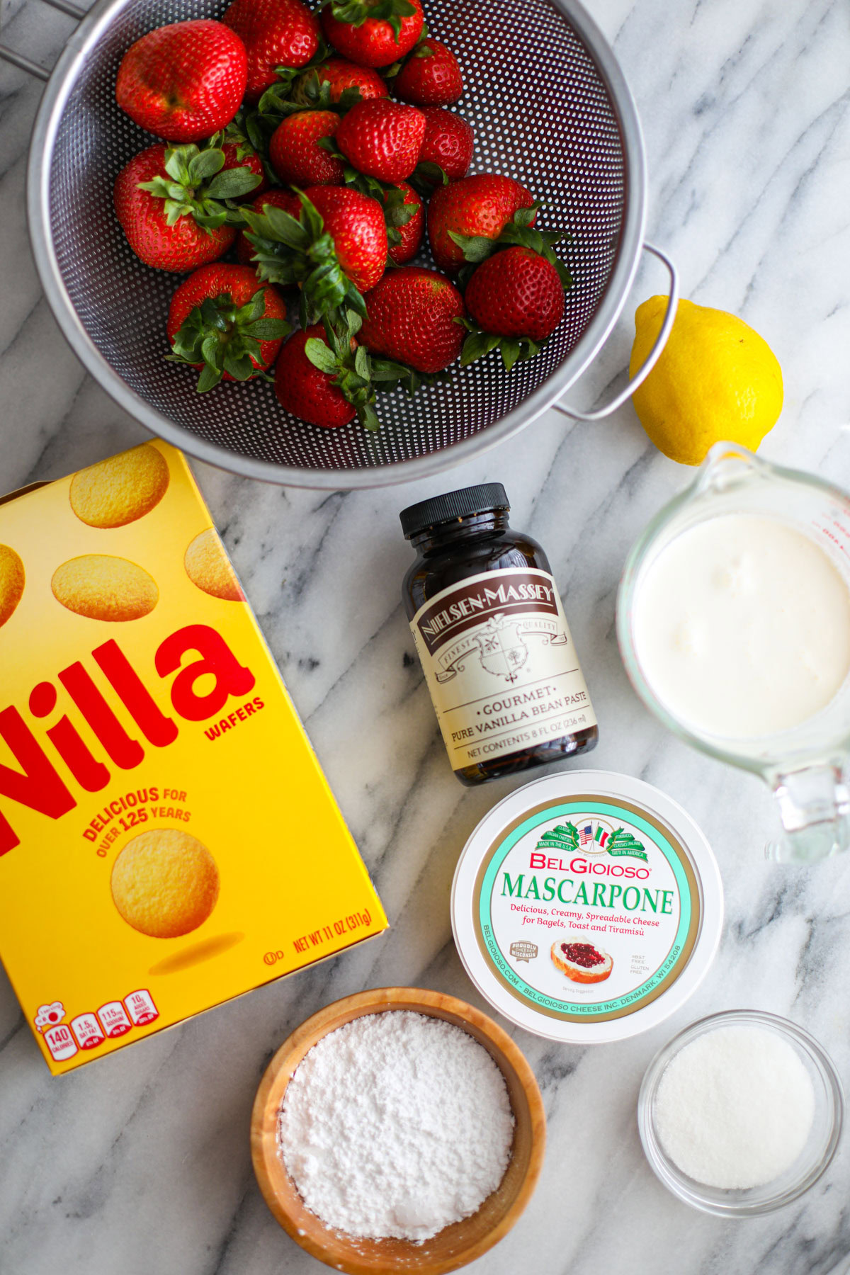 A strainer of strawberries, box of wafers, heavy cream, a lemon, vanilla bean paste, a container of mascarpone, and bowls of sugar.