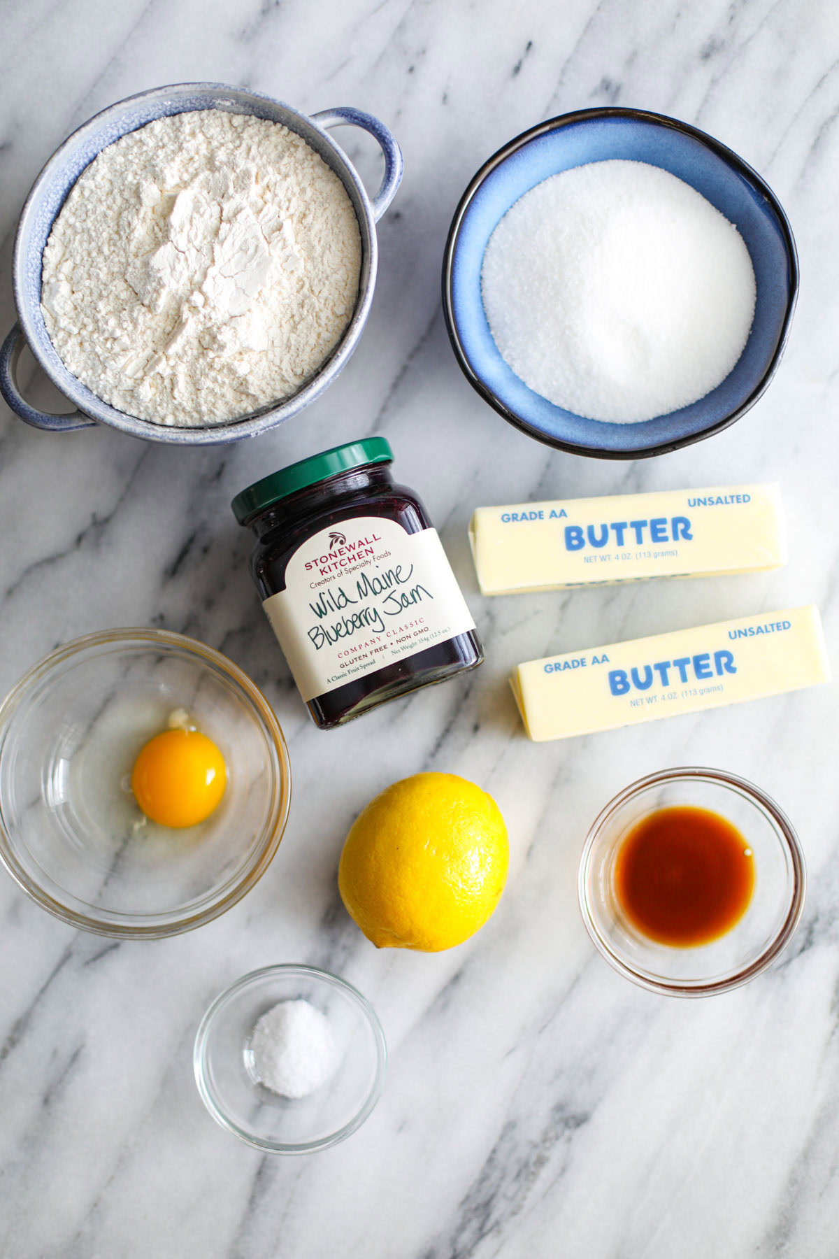 Bowls of flour, sugar, egg, vanilla, salt, two sticks of butter, a lemon, and a jar of blueberry jam.