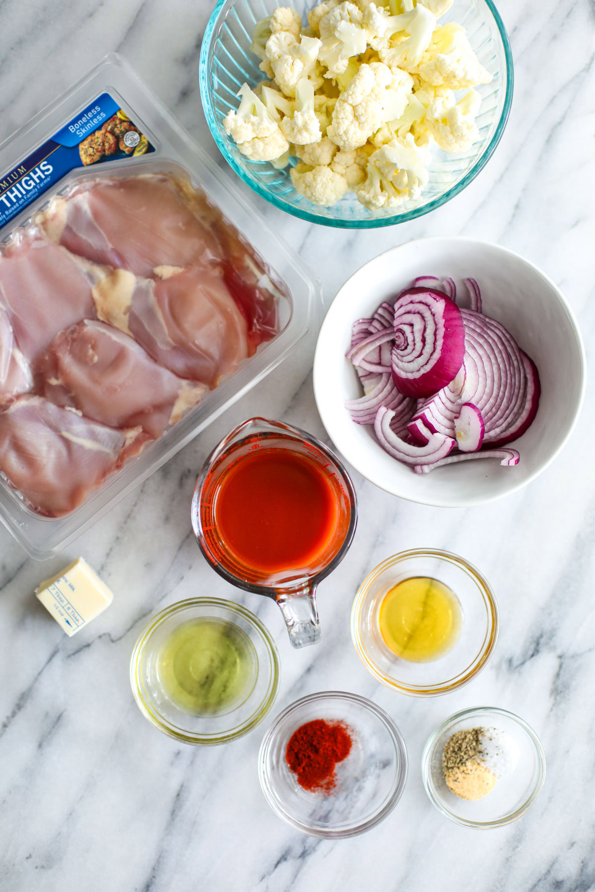 Ingredients to make buffalo chicken sheet pan dinner including chicken thighs, cauliflower, red onion, buffalo sauce, seasonings, oil, and butter.
