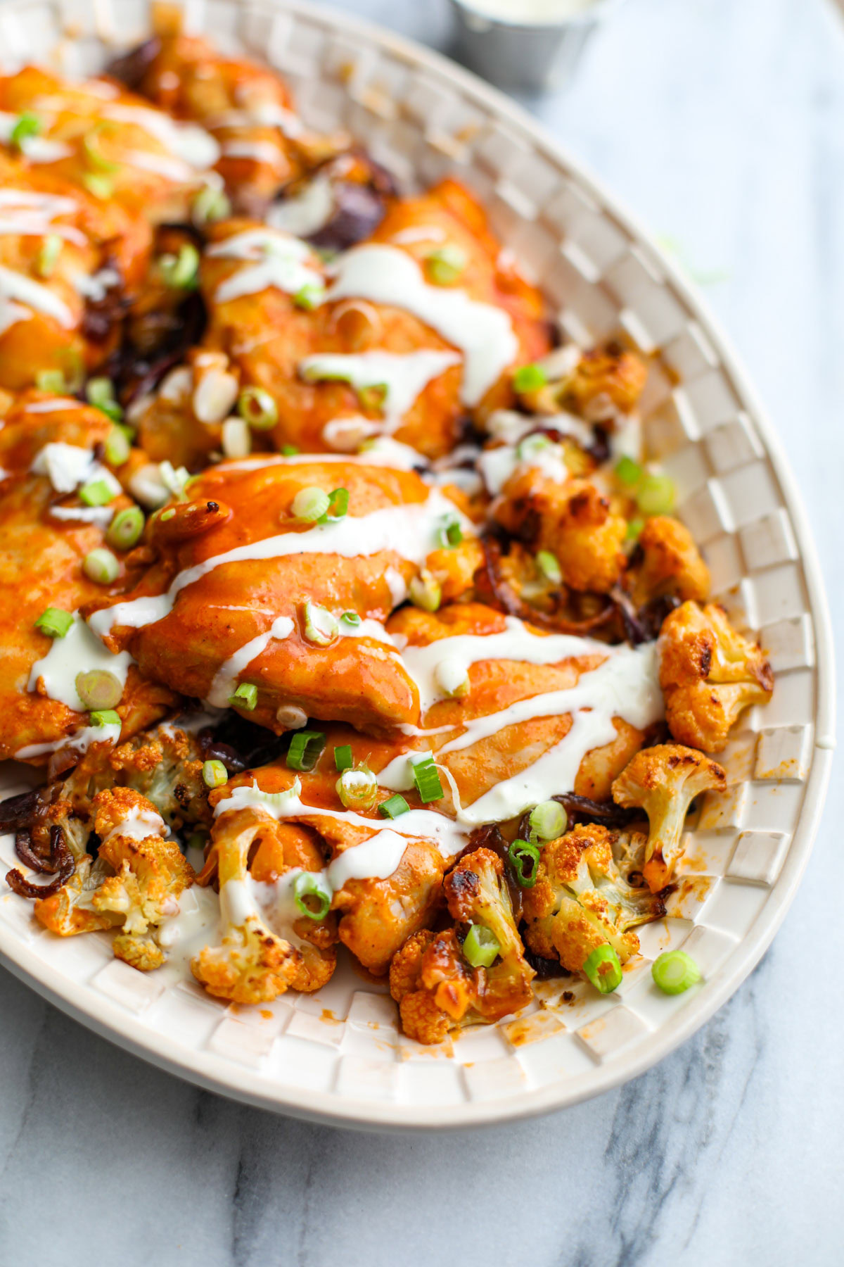 Buffalo chicken sheet pan dinner and cauliflower florets on a white platter drizzled with blue cheese and topped with green onions.