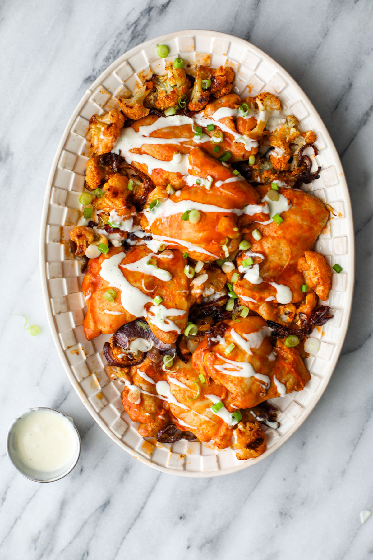 Buffalo Chicken sheet pan dinner on a white platter drizzled with blue cheese and topped with green onions and a silver condiment cup next to it.