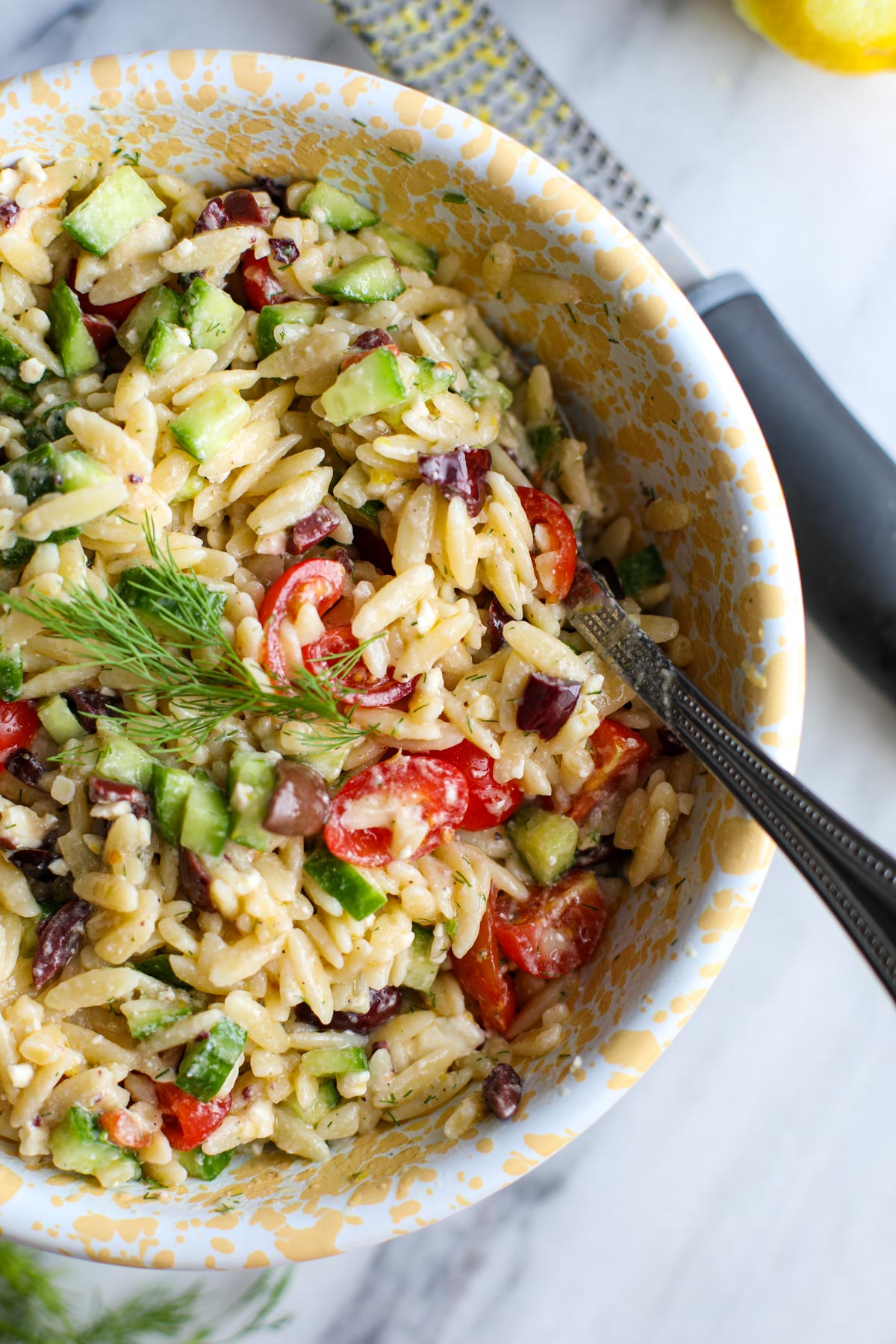 A yellow and white bowl with greek orzo pasta salad with a silver spoon.