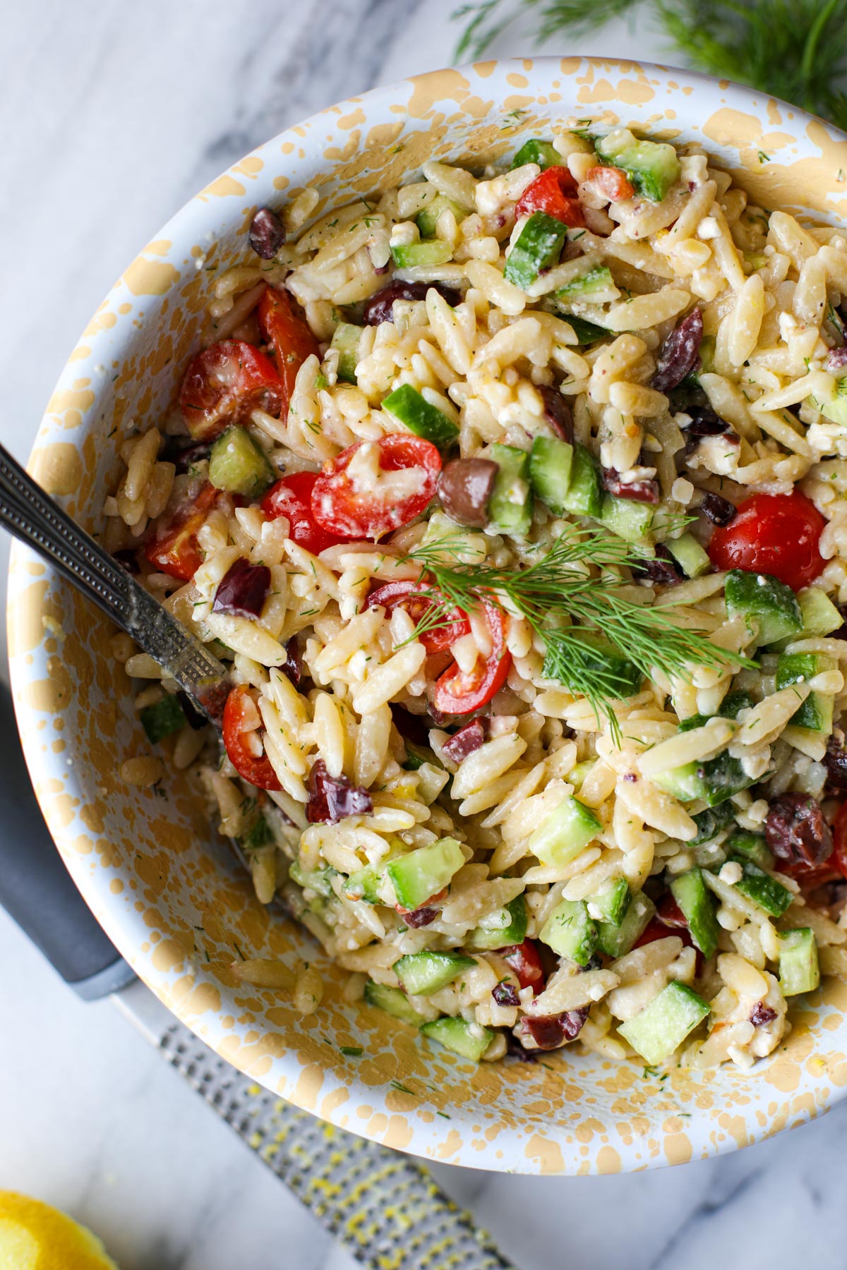 A yellow and white bowl with greek orzo pasta salad with a silver spoon.