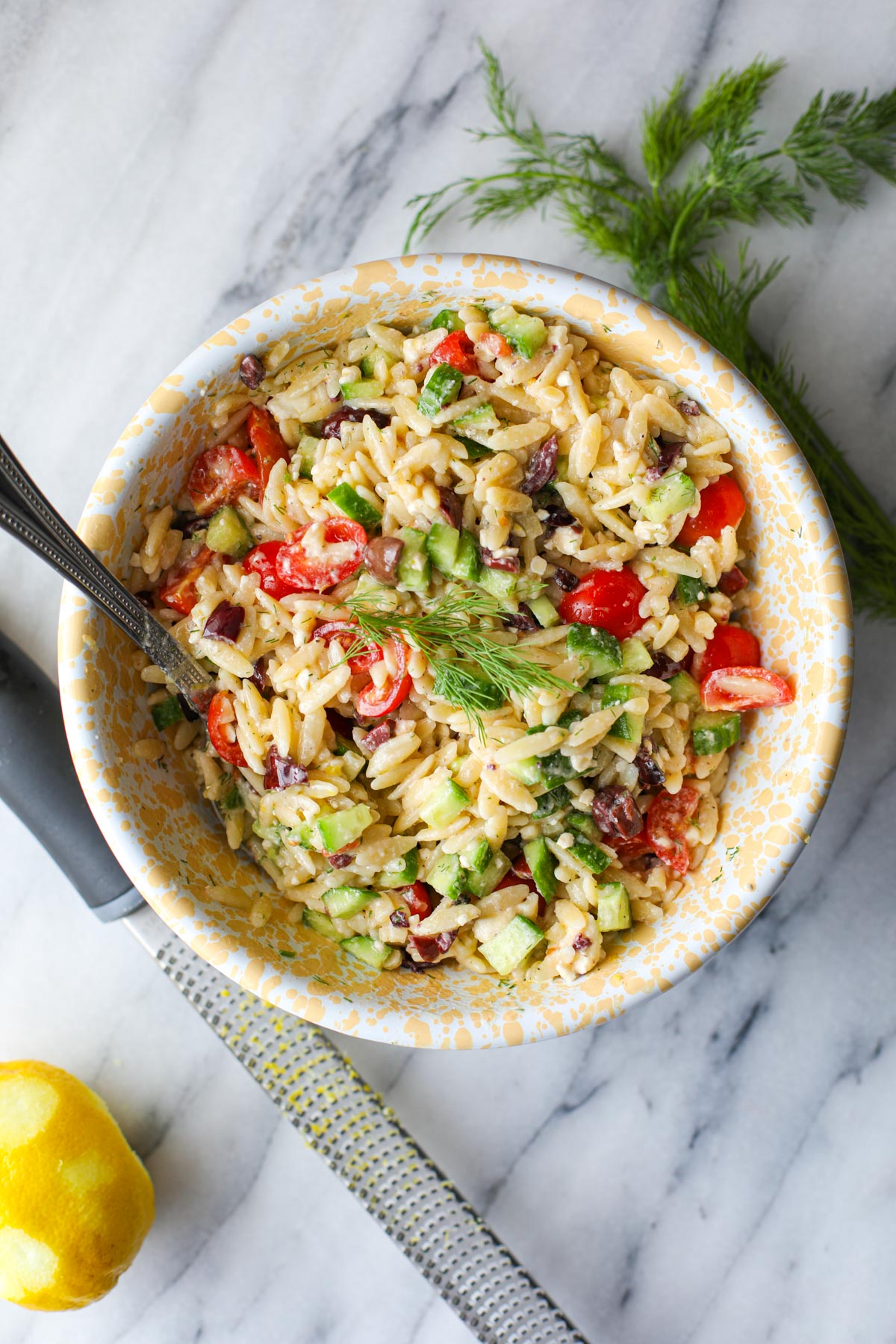 A yellow and white bowl with greek orzo pasta salad with a silver spoon next to a microplane grater and a bunch of dill.