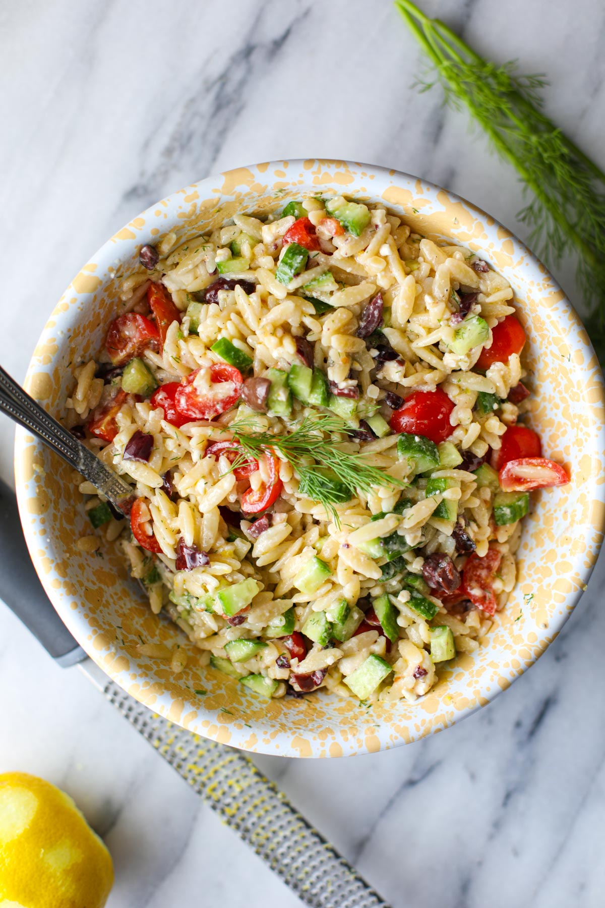 A yellow and white bowl with greek orzo pasta salad with a silver spoon.