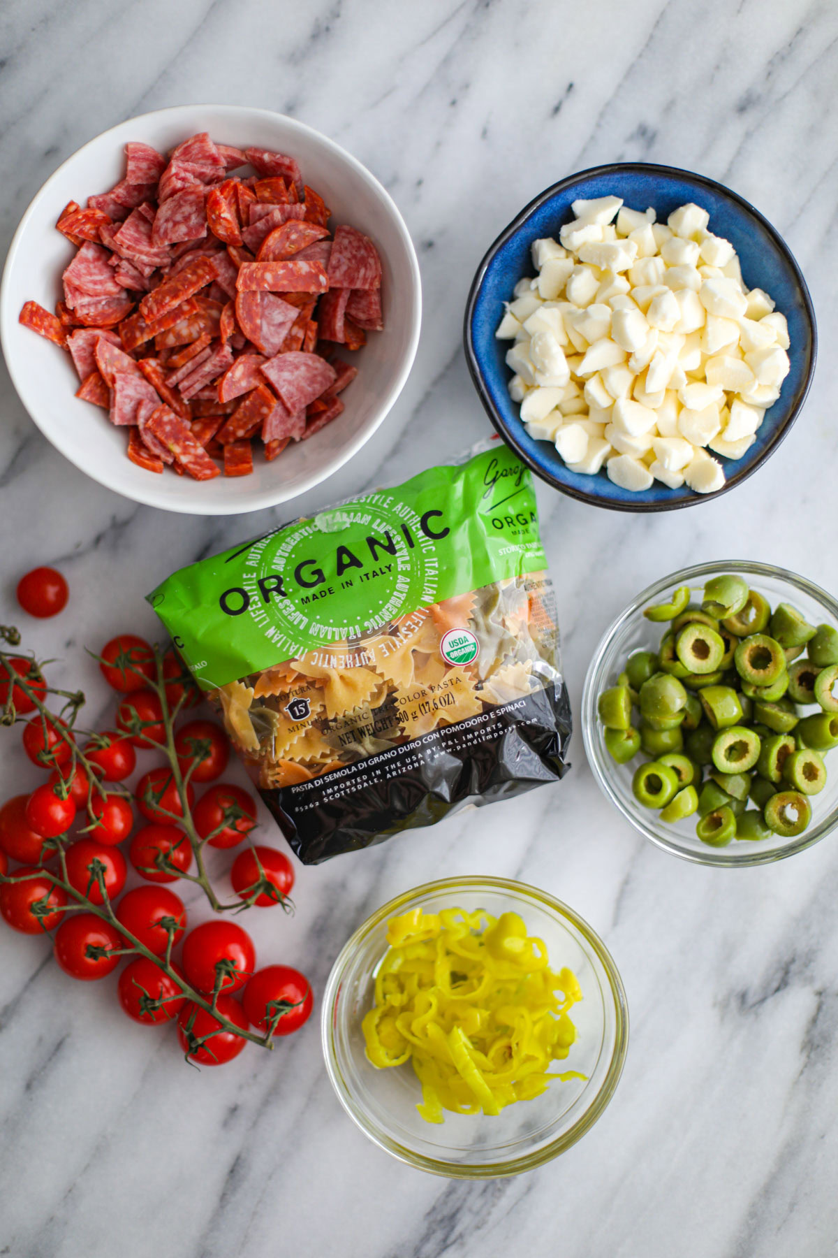 Bowls of salami, mozzarella, and olives with a bag of pasta and cherry tomatoes on the vine.