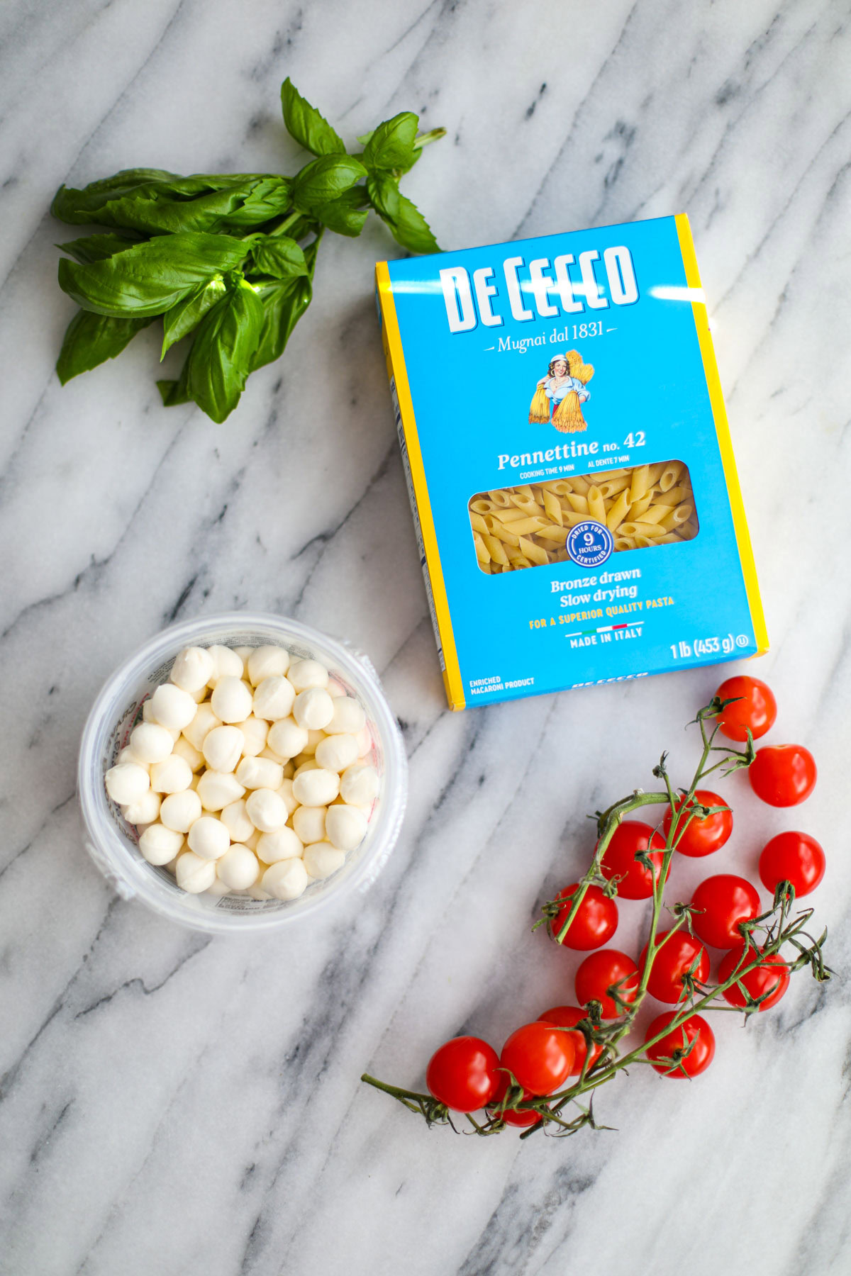 A box of pasta, a bunch of basil, cherry tomatoes on the vine, and a bowl of perline mozzarella.