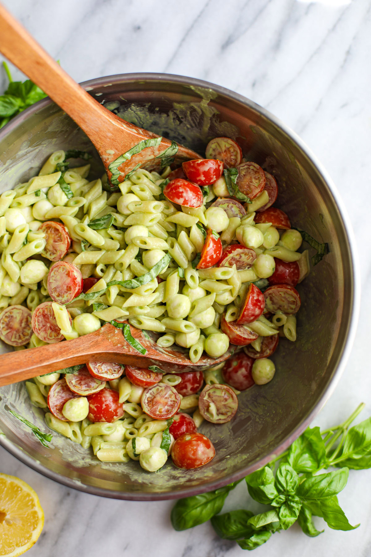 A stainless steel bowl of caprese pasta salad with wood salad servers. 