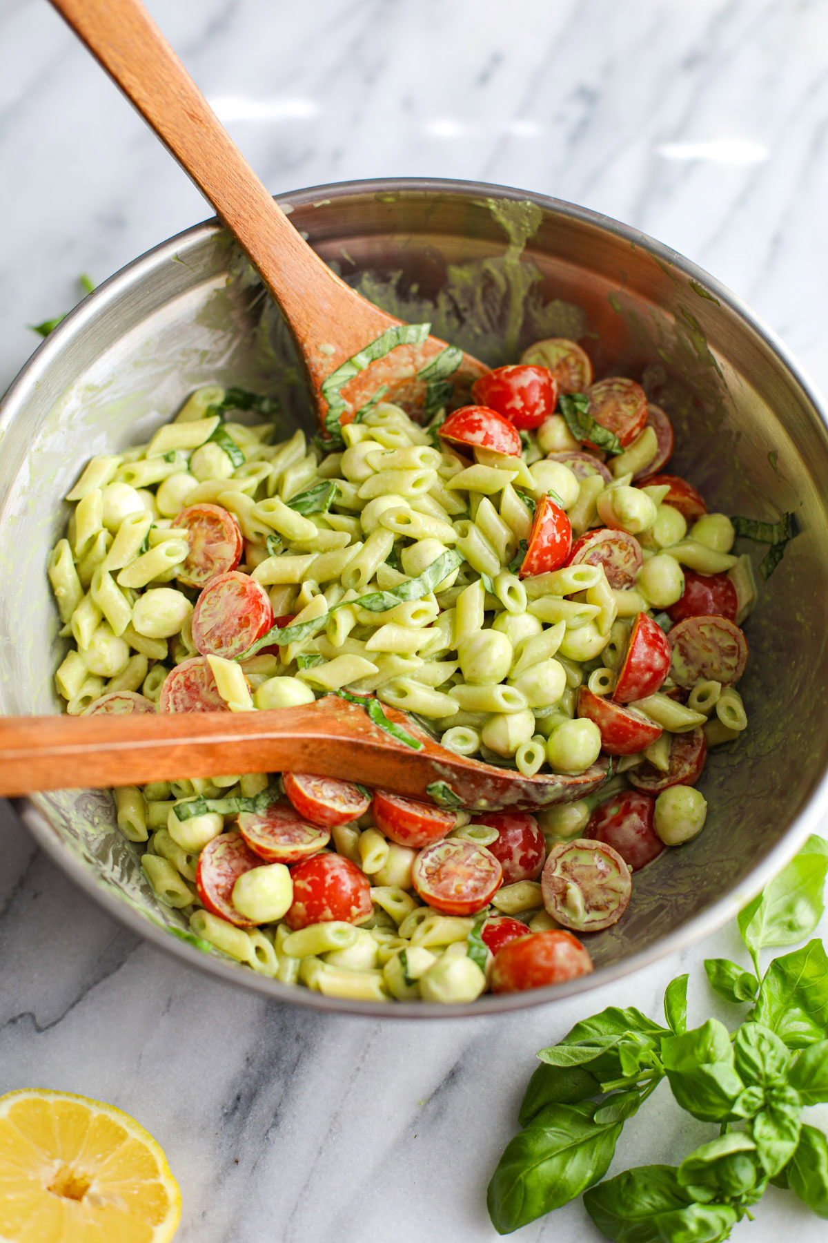 A stainless steel bowl of caprese pasta salad with wood salad servers. 